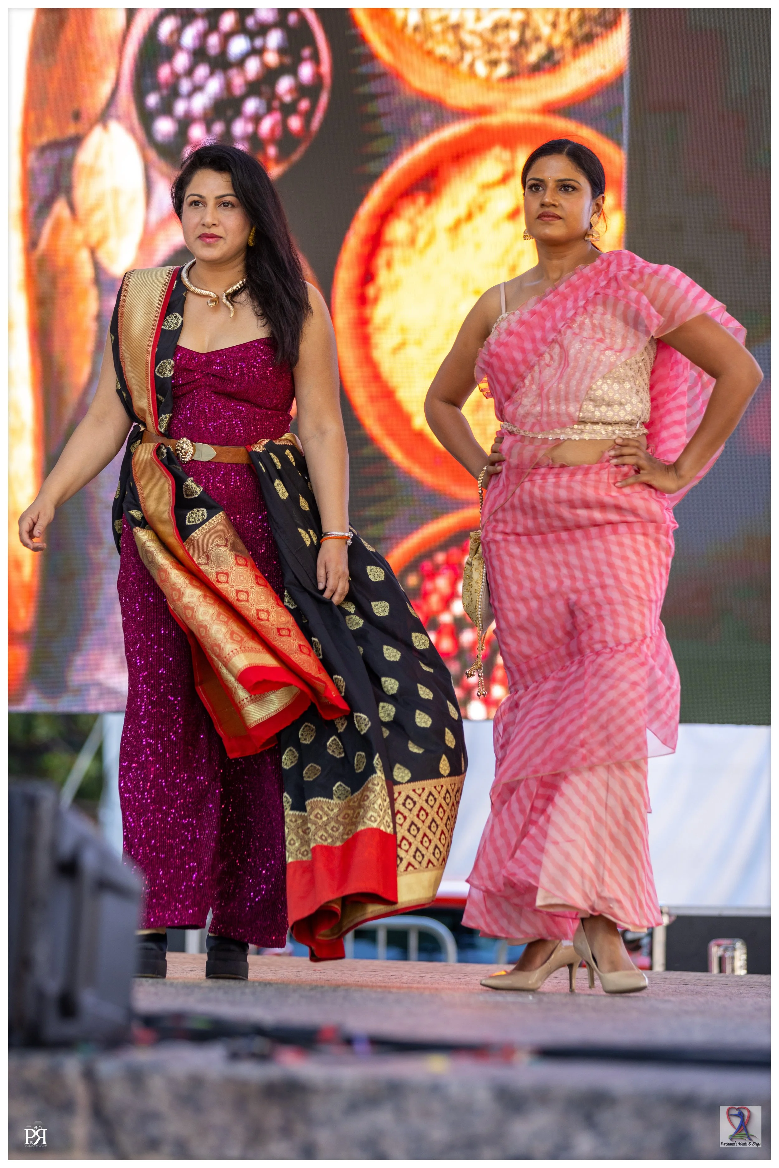 Two women dressed in colorful traditional Indian clothing, standing on stage with a background display of fruit images.
