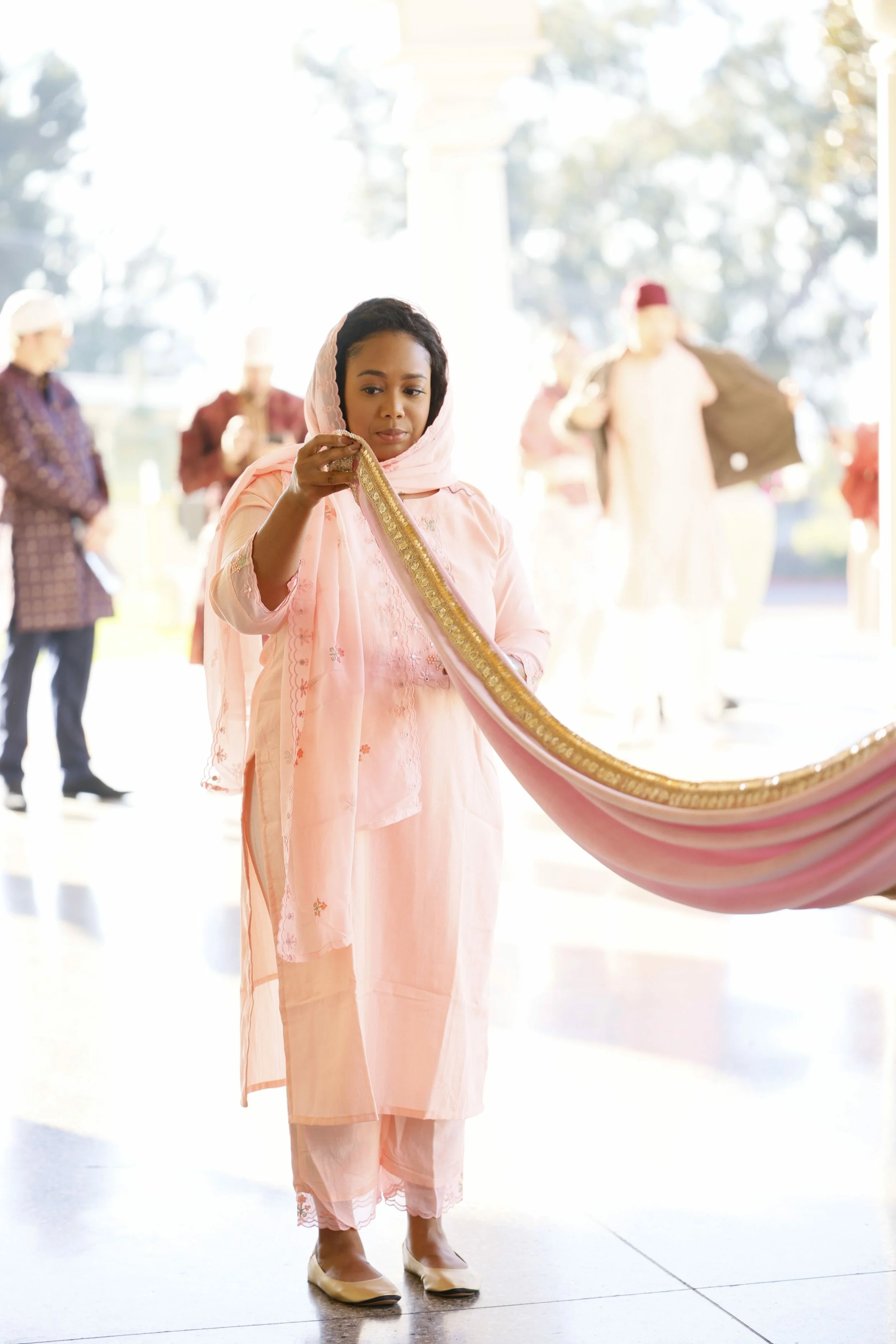 Woman in a pink traditional outfit holding a draped pink and gold fabric, with other people in traditional clothing in the background.