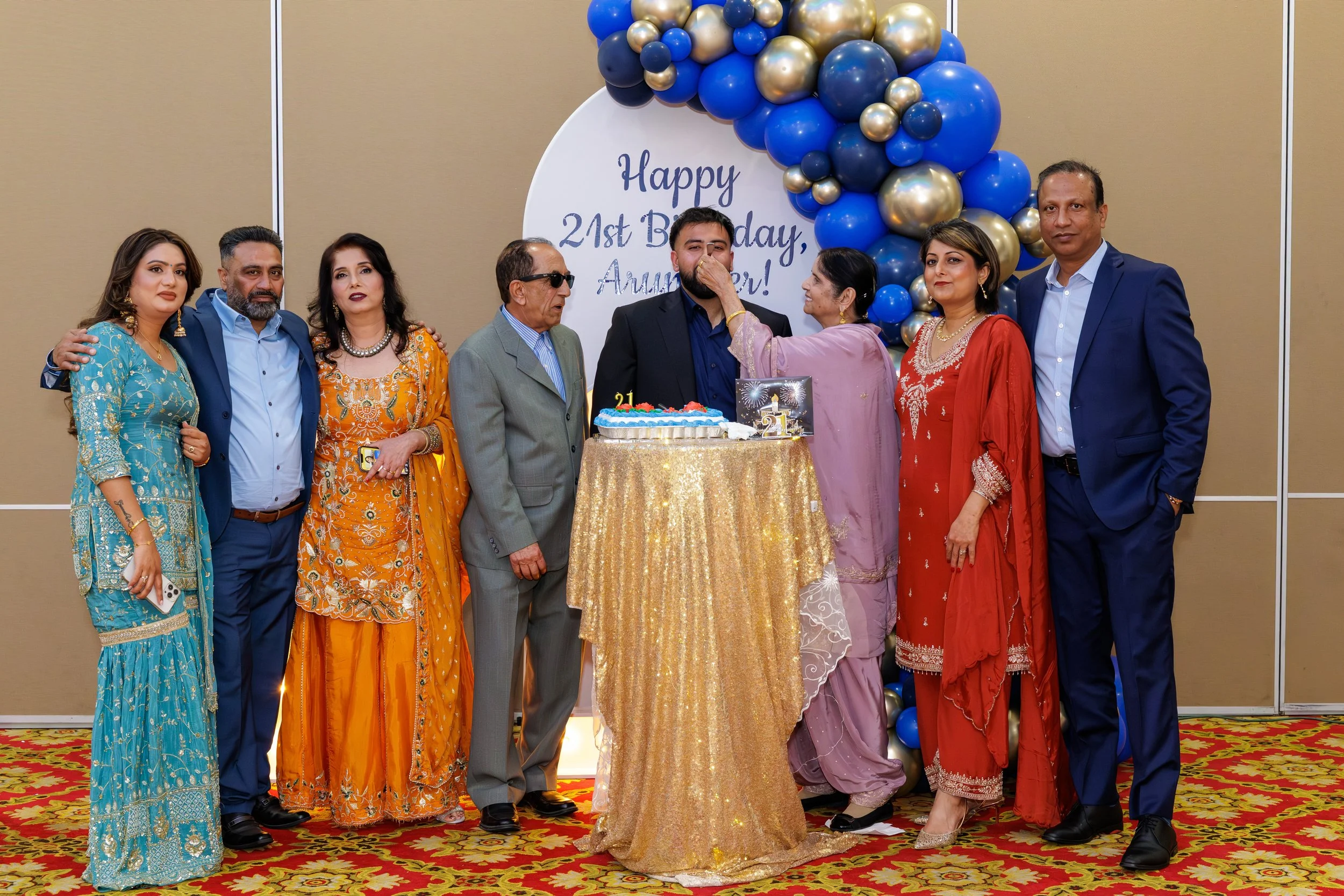 A group of nine people celebrating a birthday, with a decorated backdrop, balloons, a cake, and a woman feeding a man a piece of cake.