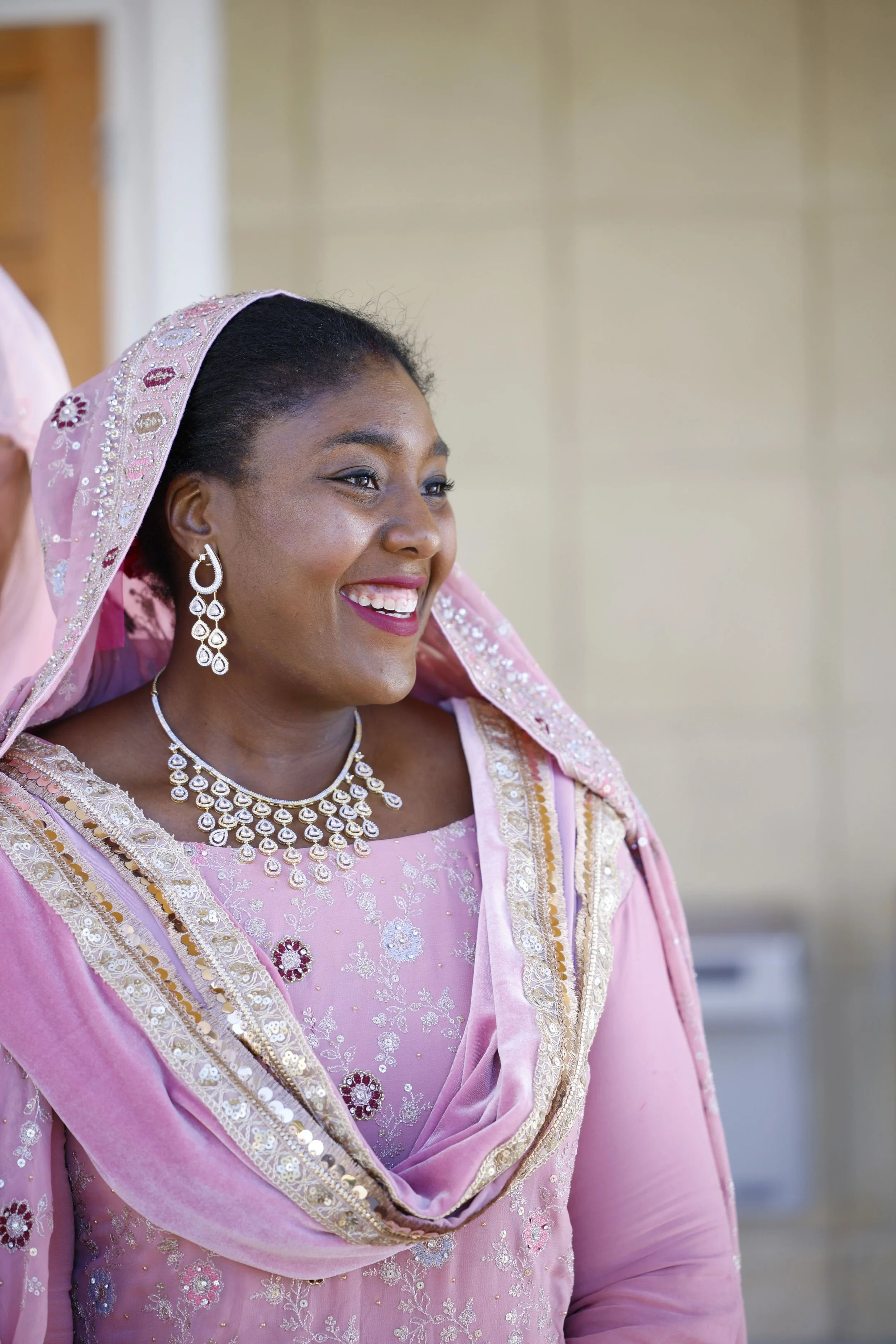 A woman wearing a pink embroidered dress with matching headscarf, jewelry, and earrings, smiling.