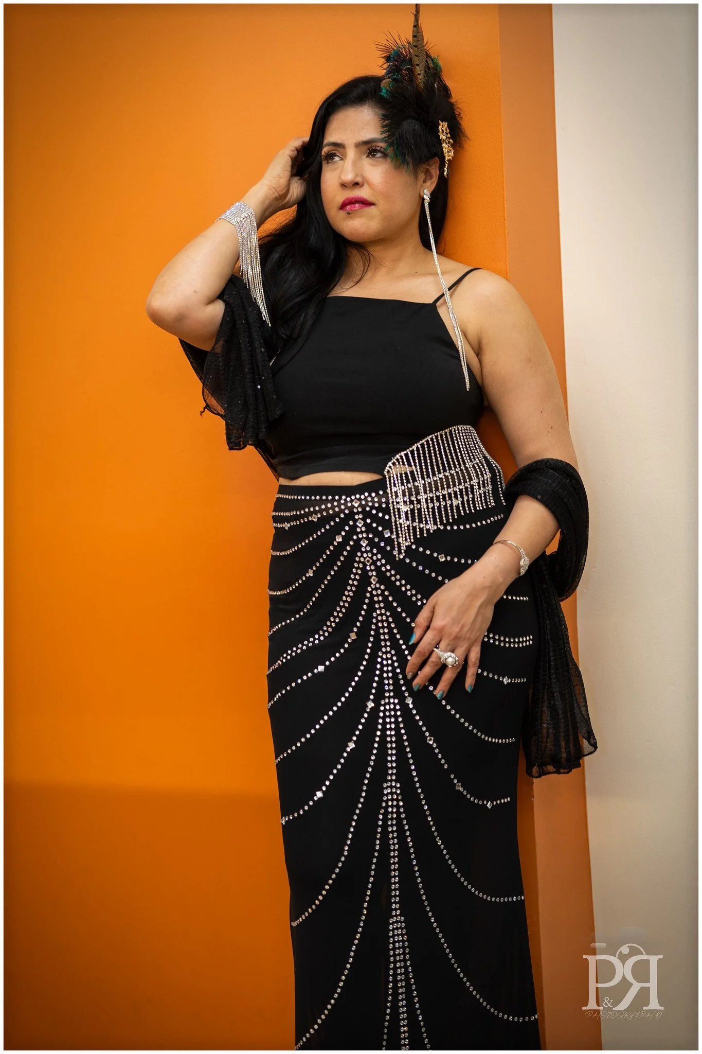 Woman in black dress with rhinestone embellishments, standing against orange and white walls, wearing a feathered headpiece and jewelry.