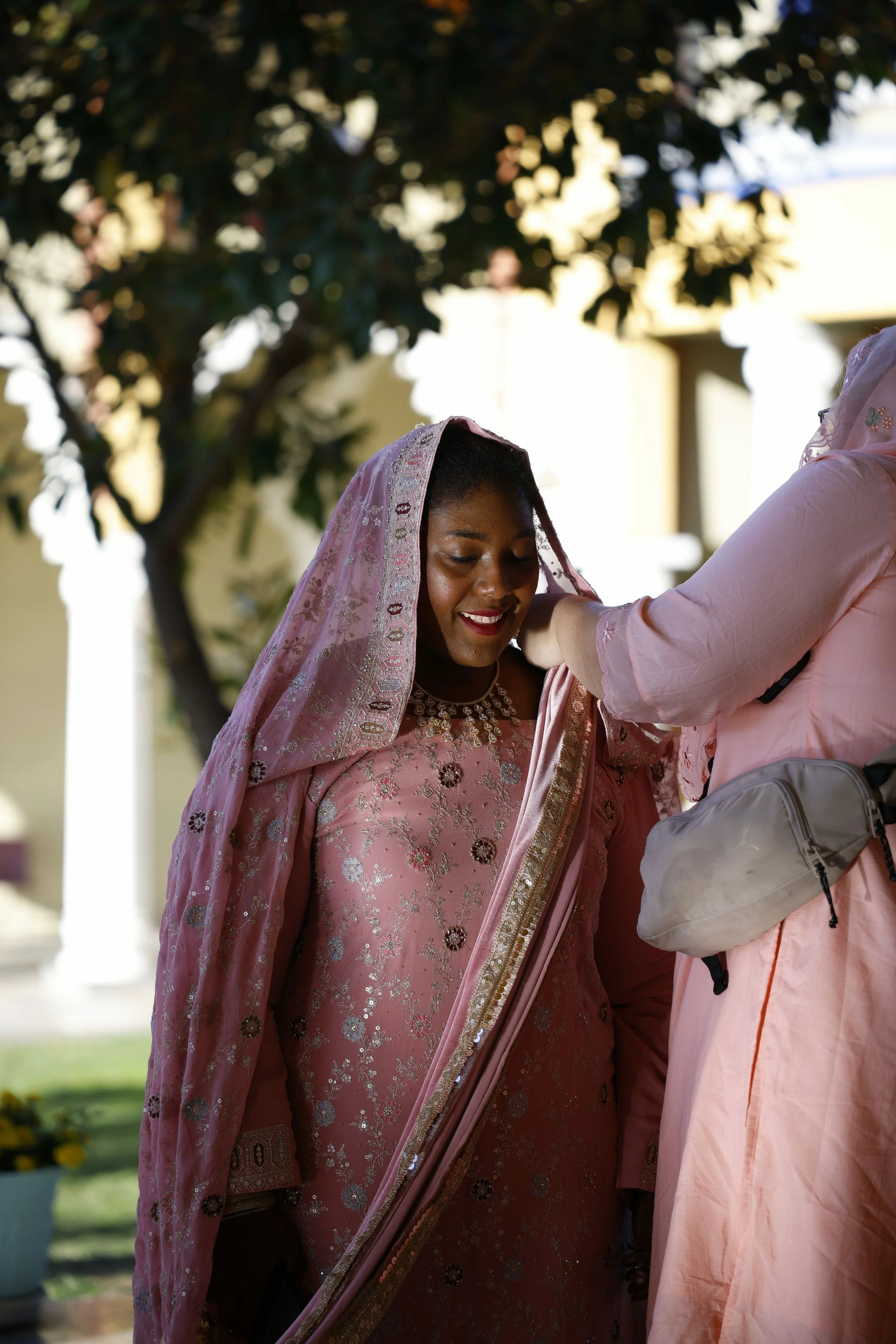 A woman dressed in traditional pink Indian attire smiling as someone adjusts her outfit outdoors, with a tree and building in the background.