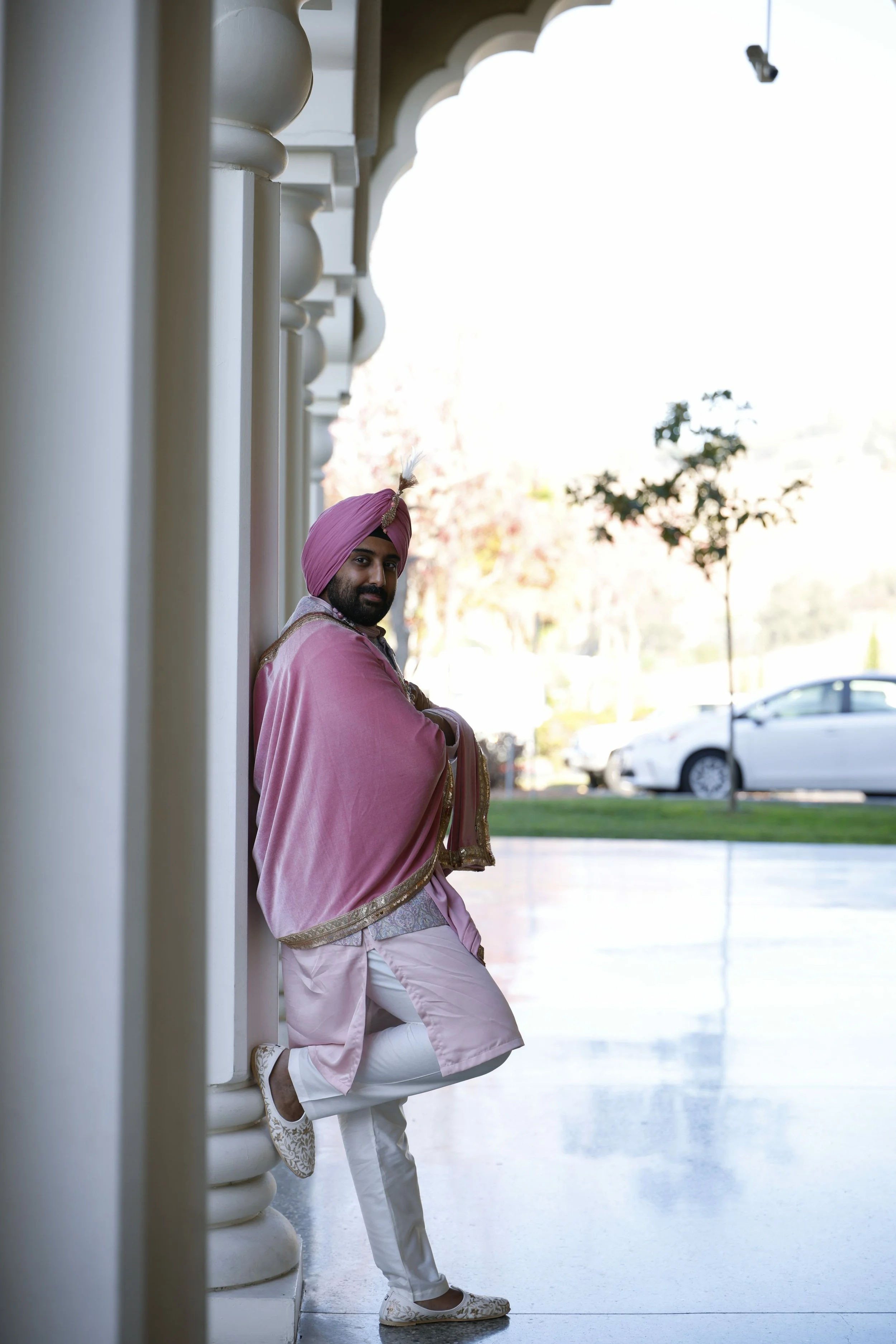 A man dressed in traditional pink and white Indian attire, wearing a pink turban with a feather, standing against a white columned wall outside a building, with cars and trees in the background.