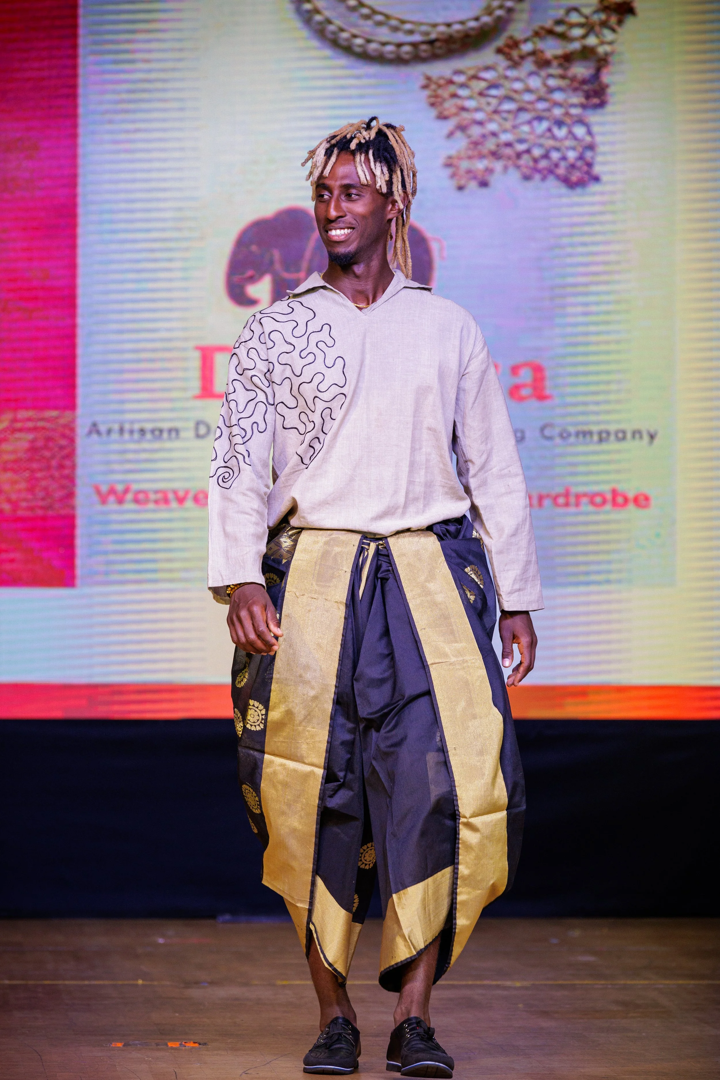 A man walking on a runway wearing a traditional African style outfit with black and yellow fabric, a long shirt with pattern, and black shoes. A colorful background with text and images is behind him.