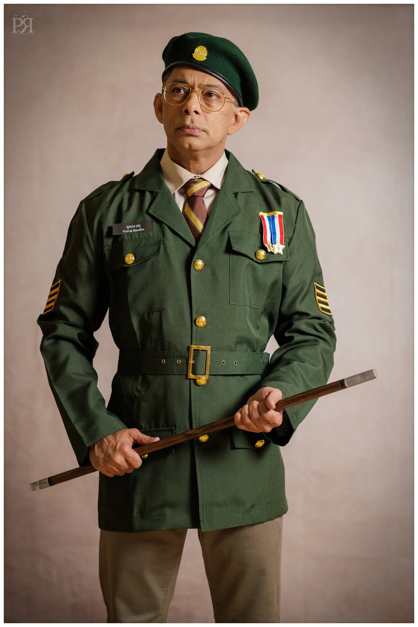 A man dressed in a green military uniform, holding a wooden stick, wearing glasses, and a beret with a badge, standing against a plain background.