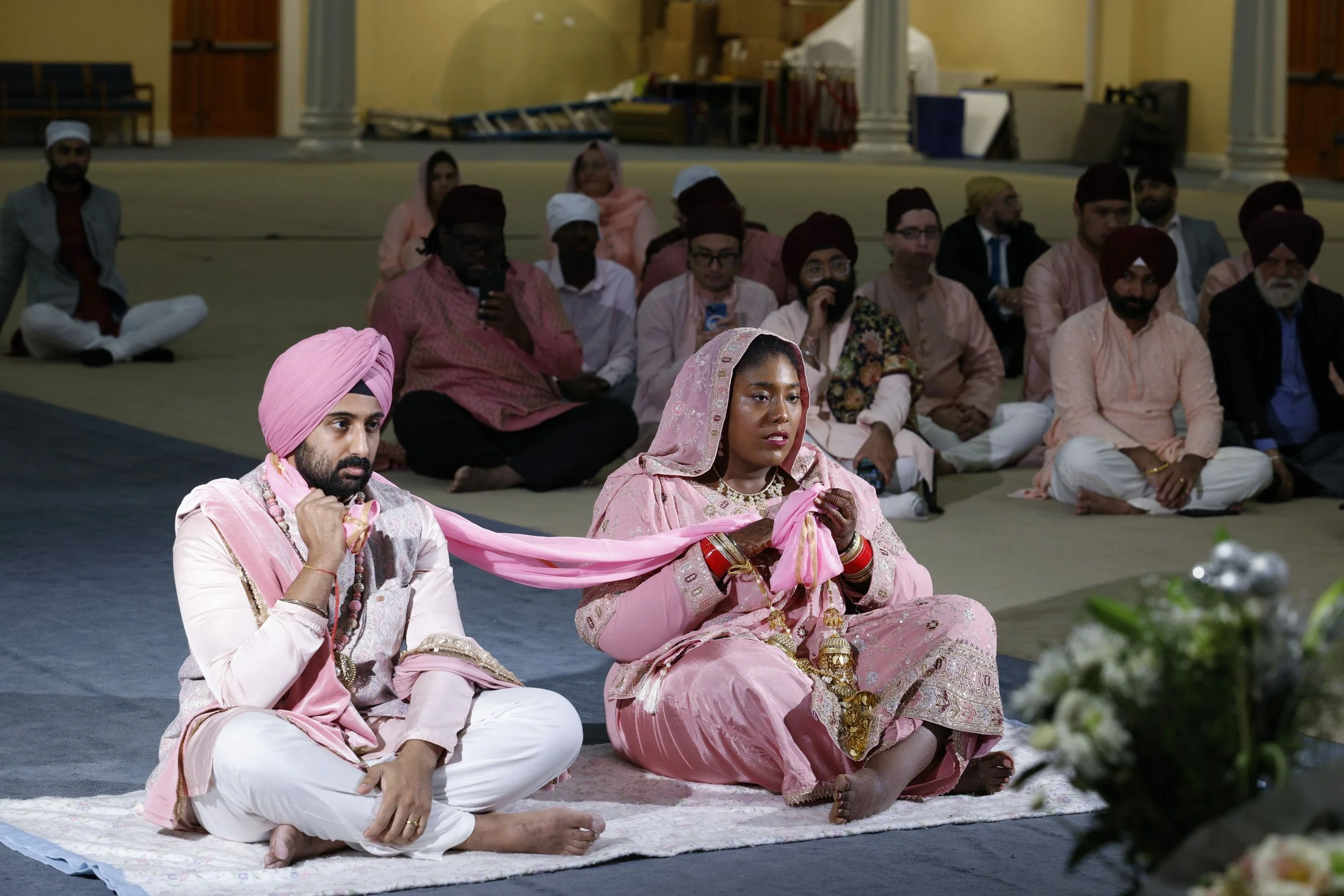 Indian couple in traditional wedding attire, sitting on the floor with a pink scarf, during a wedding ceremony with guests seated behind them.