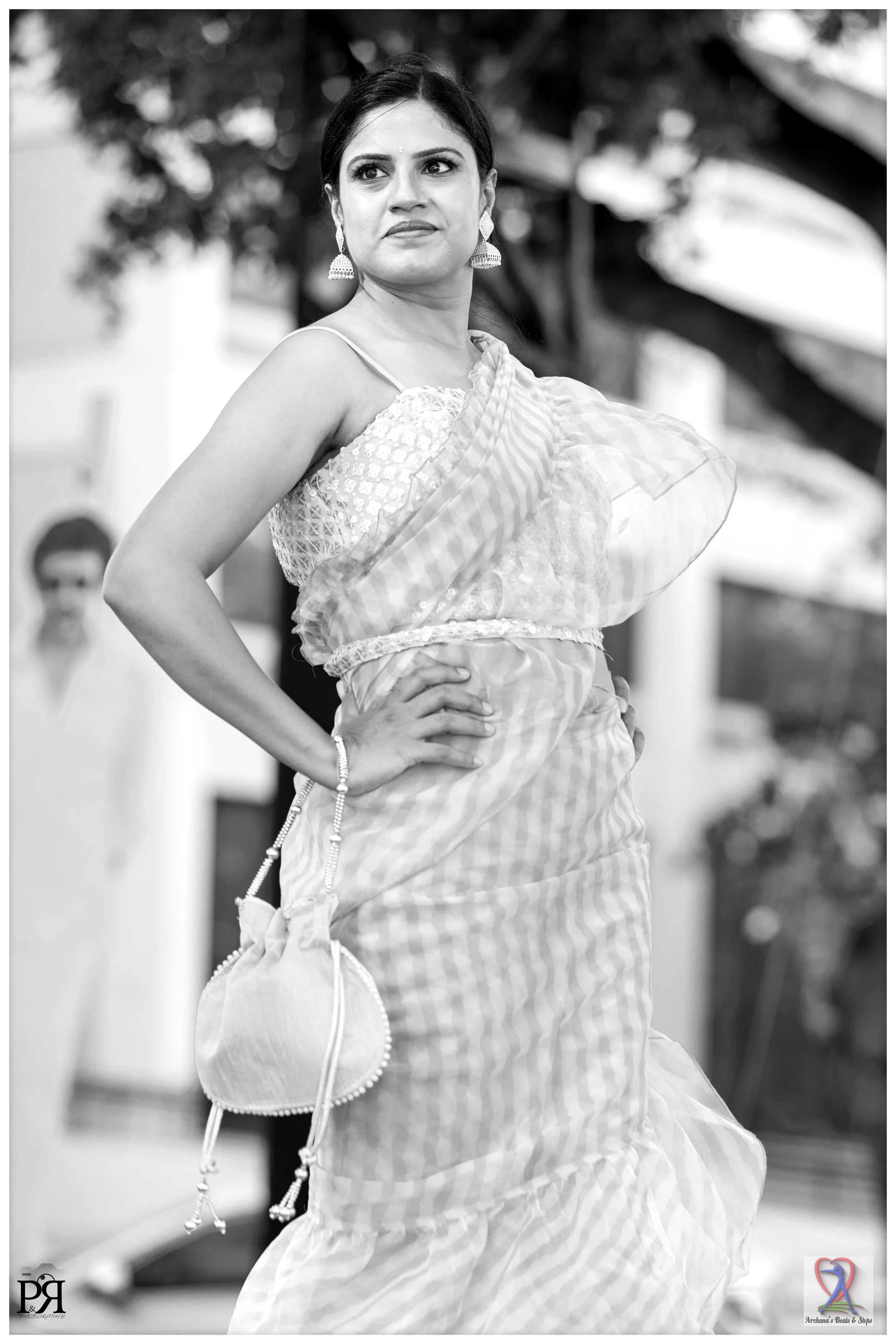 A woman in traditional Indian attire, wearing earrings and a sari, standing outdoors with a confident stance, hands on her hips, in black and white photo.