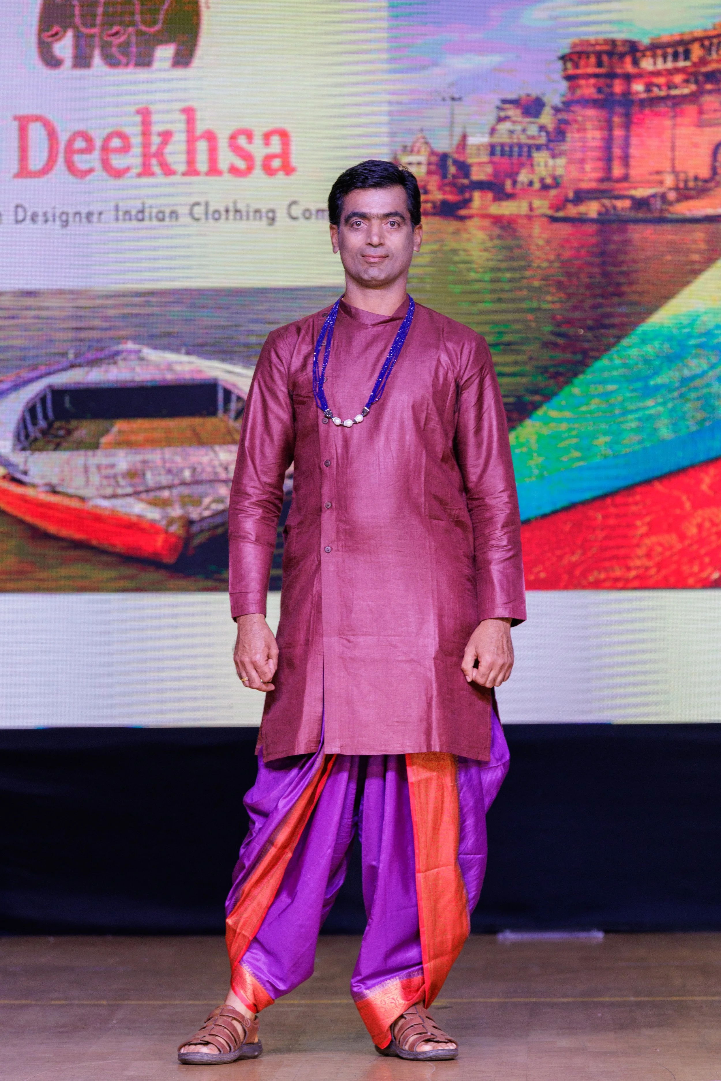 A man wearing traditional Indian attire—maroon kurta and purple dhoti with orange accents—standing on stage in front of a large digital screen displaying images of a boat, cityscape, and colorful design, with the text 'Dekhsa' and 'Indian Clothing'.