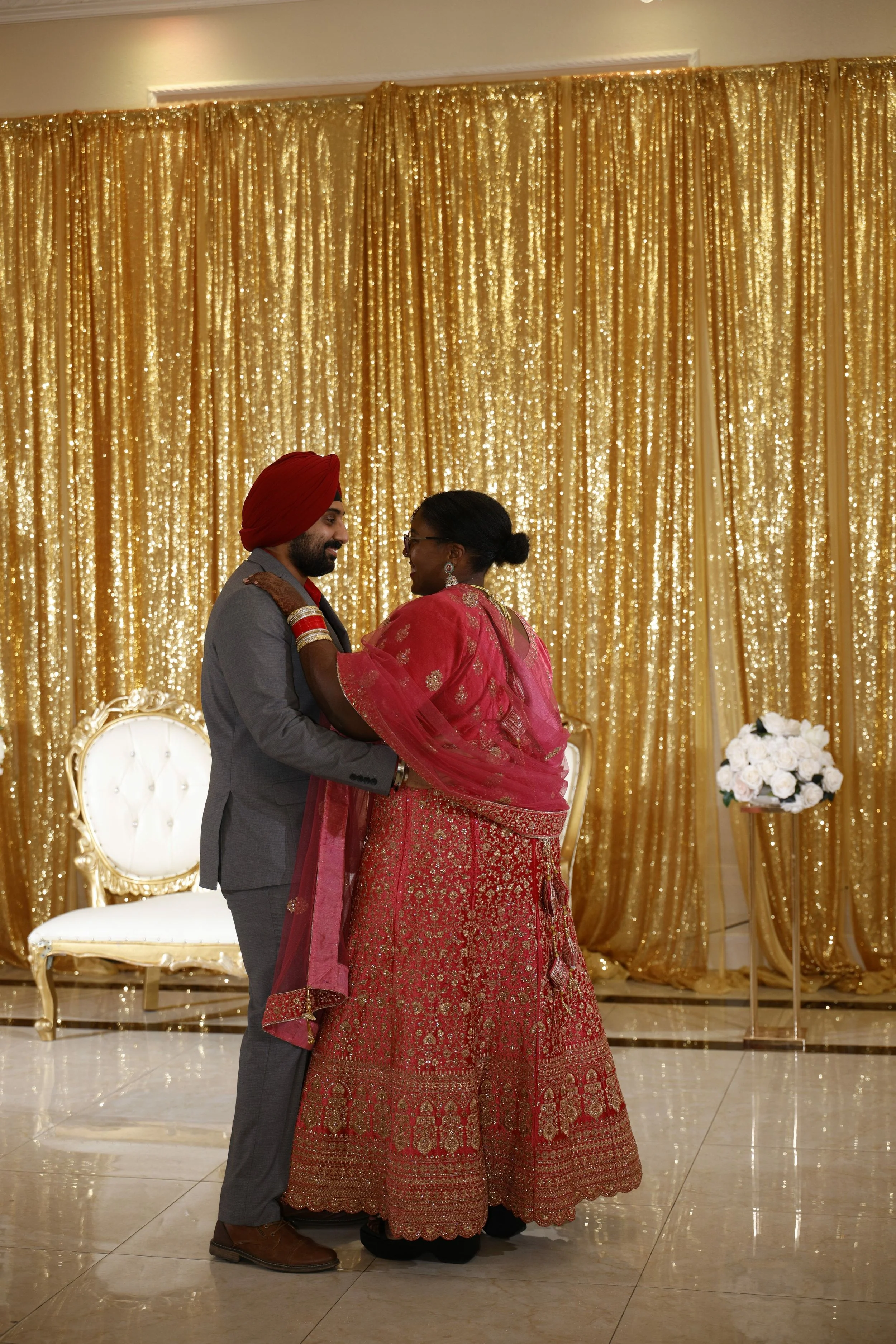 A couple dancing during a celebration, with the man wearing a gray suit and red turban, and the woman in a red traditional dress, against a background of gold glittering curtains and white chairs.