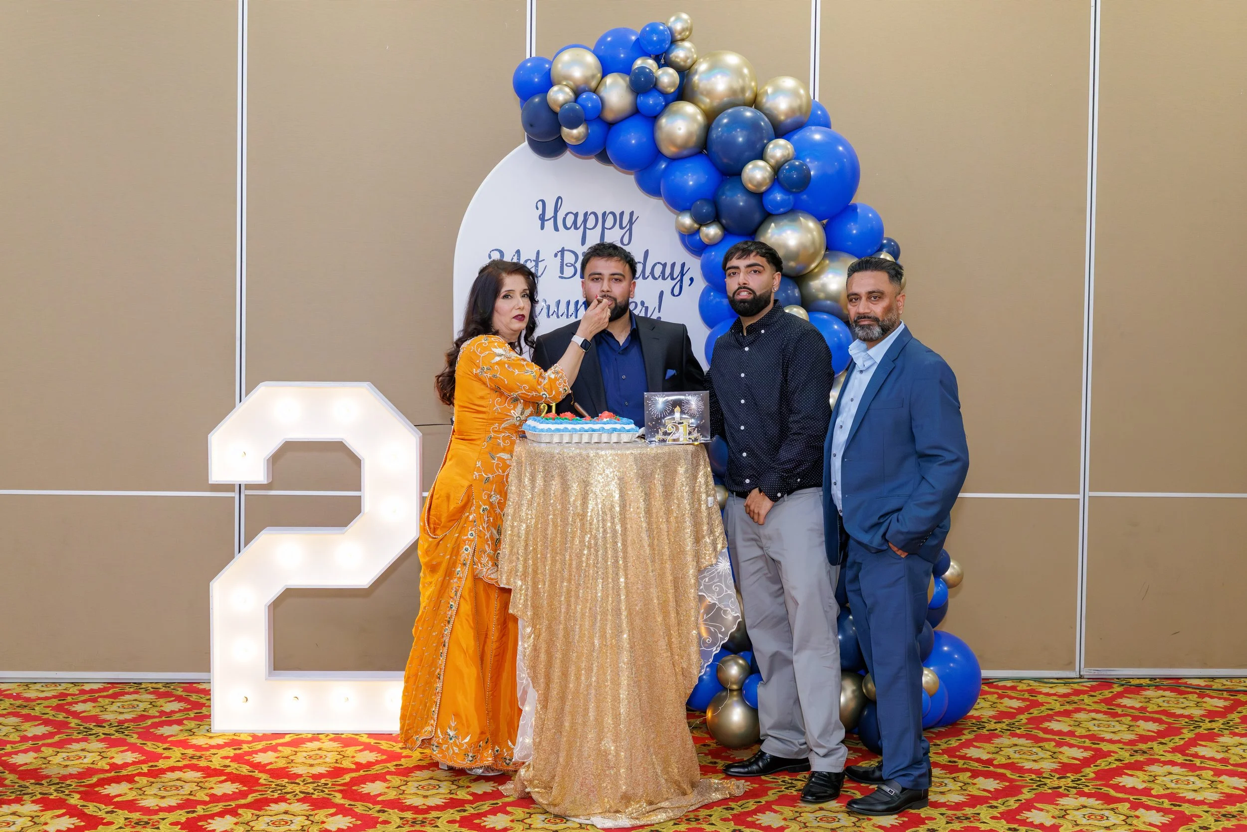 Family celebrating a birthday with a decorated balloon arch and a cake, standing in front of a sign that reads 'Happy 21st Birthday, [Name]!' and a large illuminated number 2.
