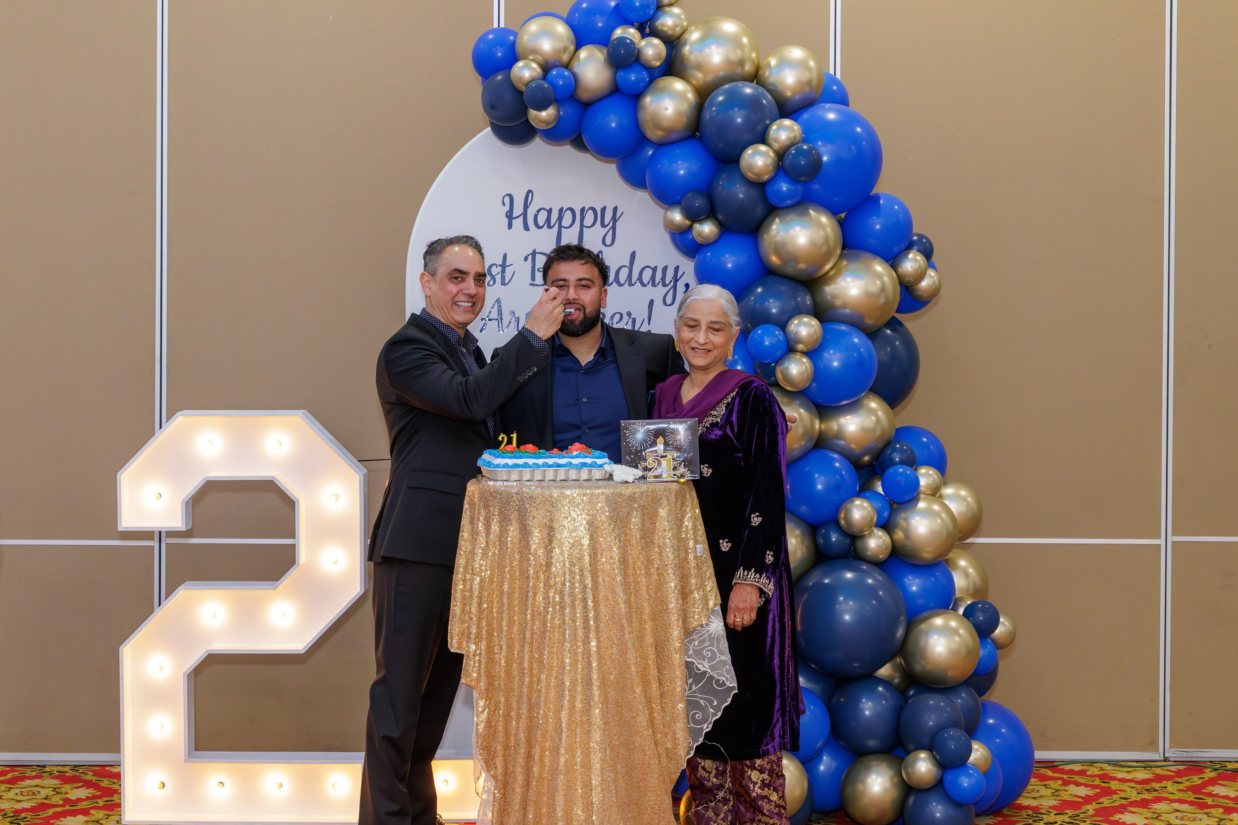 A family celebrating a 21st birthday with a cake, surrounded by blue and gold balloons, and a large illuminated number 2. A sign in the background reads "Happy 21st Birthday, Armen!"