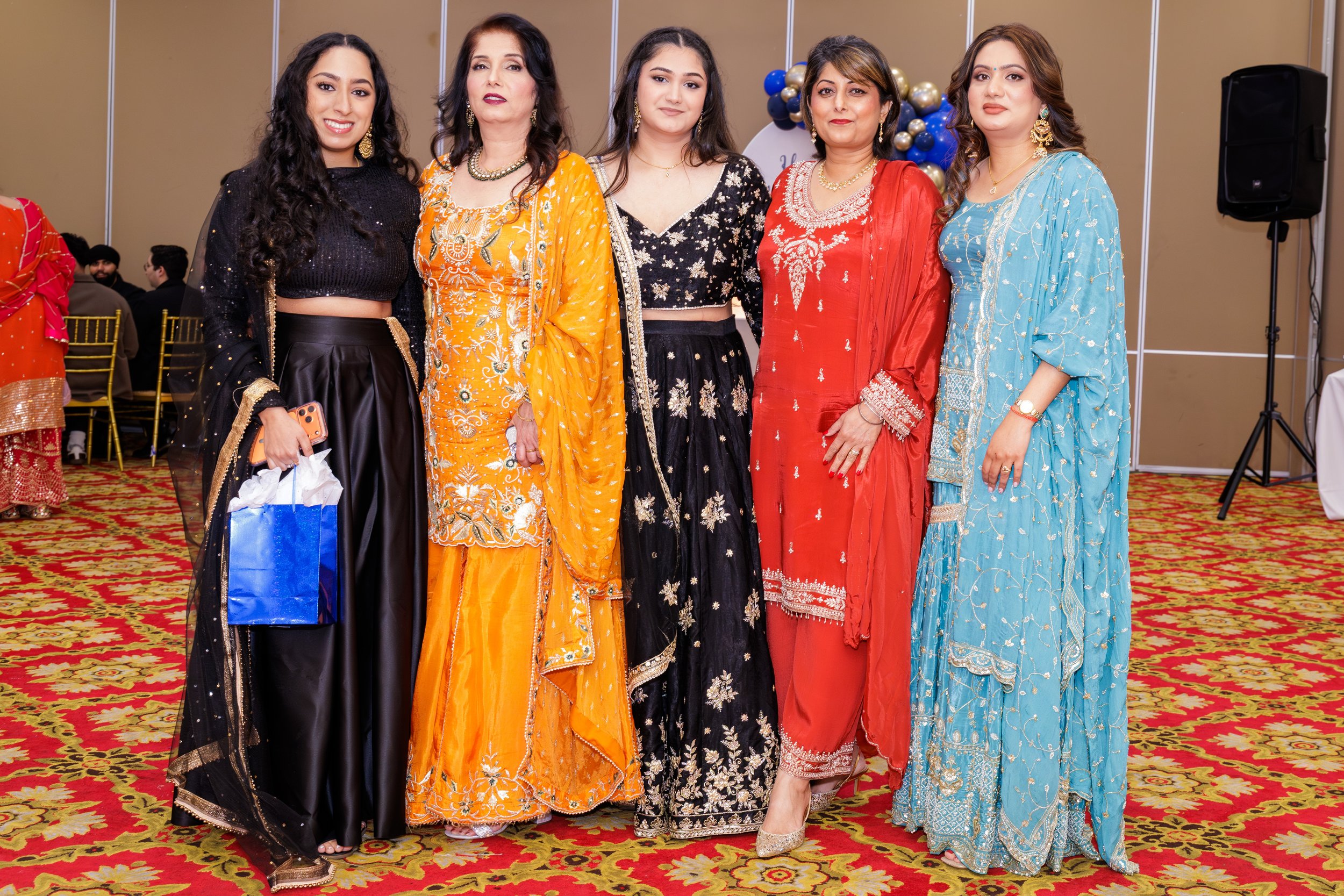 Group of six women dressed in colorful traditional South Asian attire standing indoors on a patterned carpet with balloons in the background.