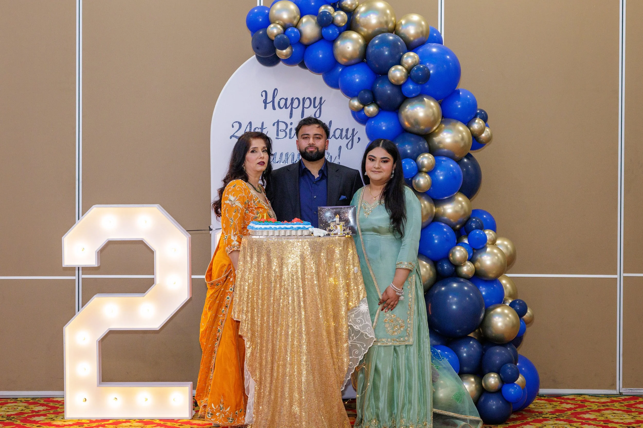 Three people standing around a table with a birthday cake, celebrating a 21st birthday. Behind them is a balloon arch in blue, gold, and navy, with a large illuminated number 2 on the left and a sign in the background reading "Happy 21st Birthday, (n