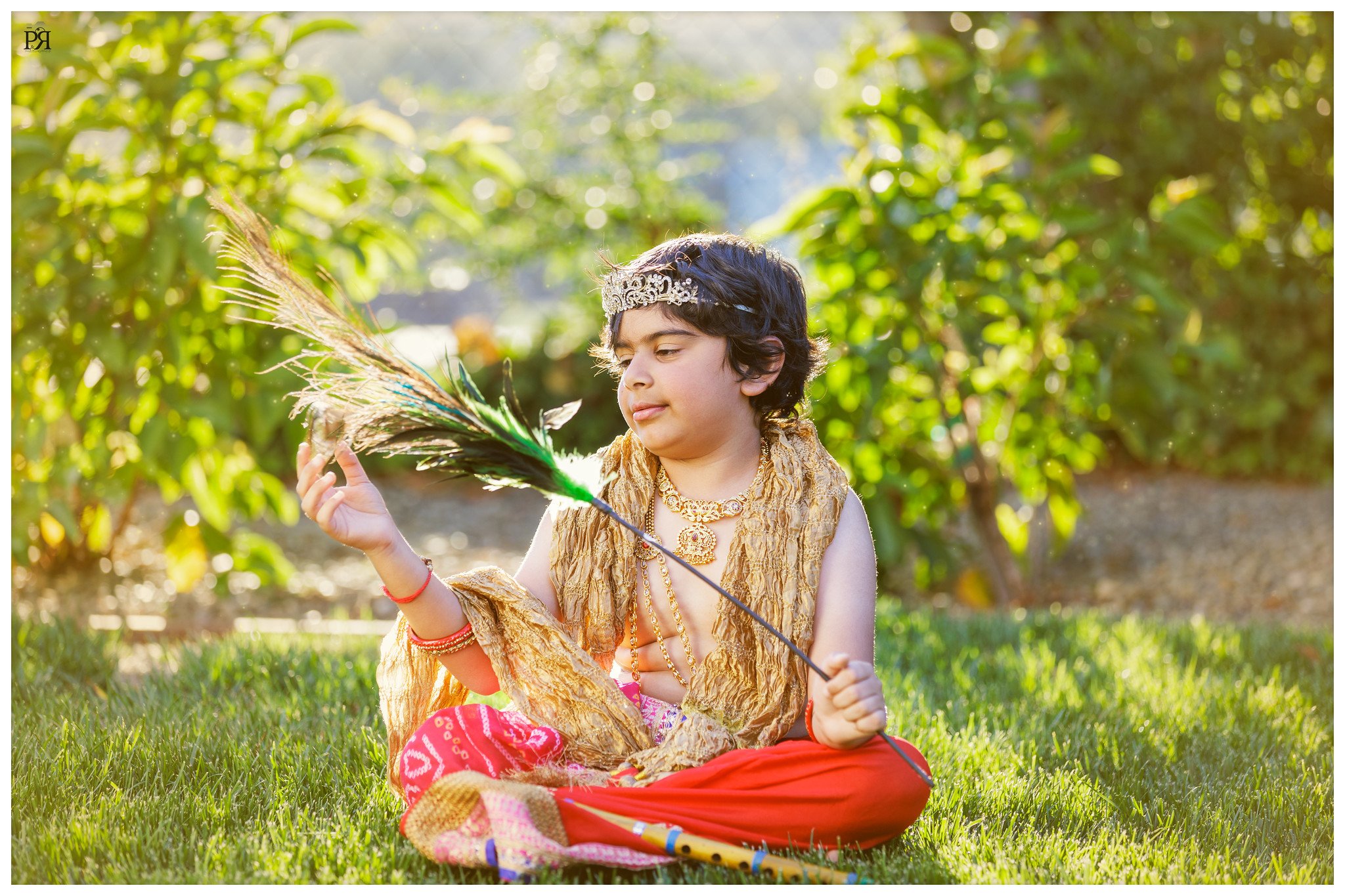 A young boy dressed in traditional Indian attire, wearing a crown and jewelry, is sitting on the grass and holding a feathered arrow while looking at it.