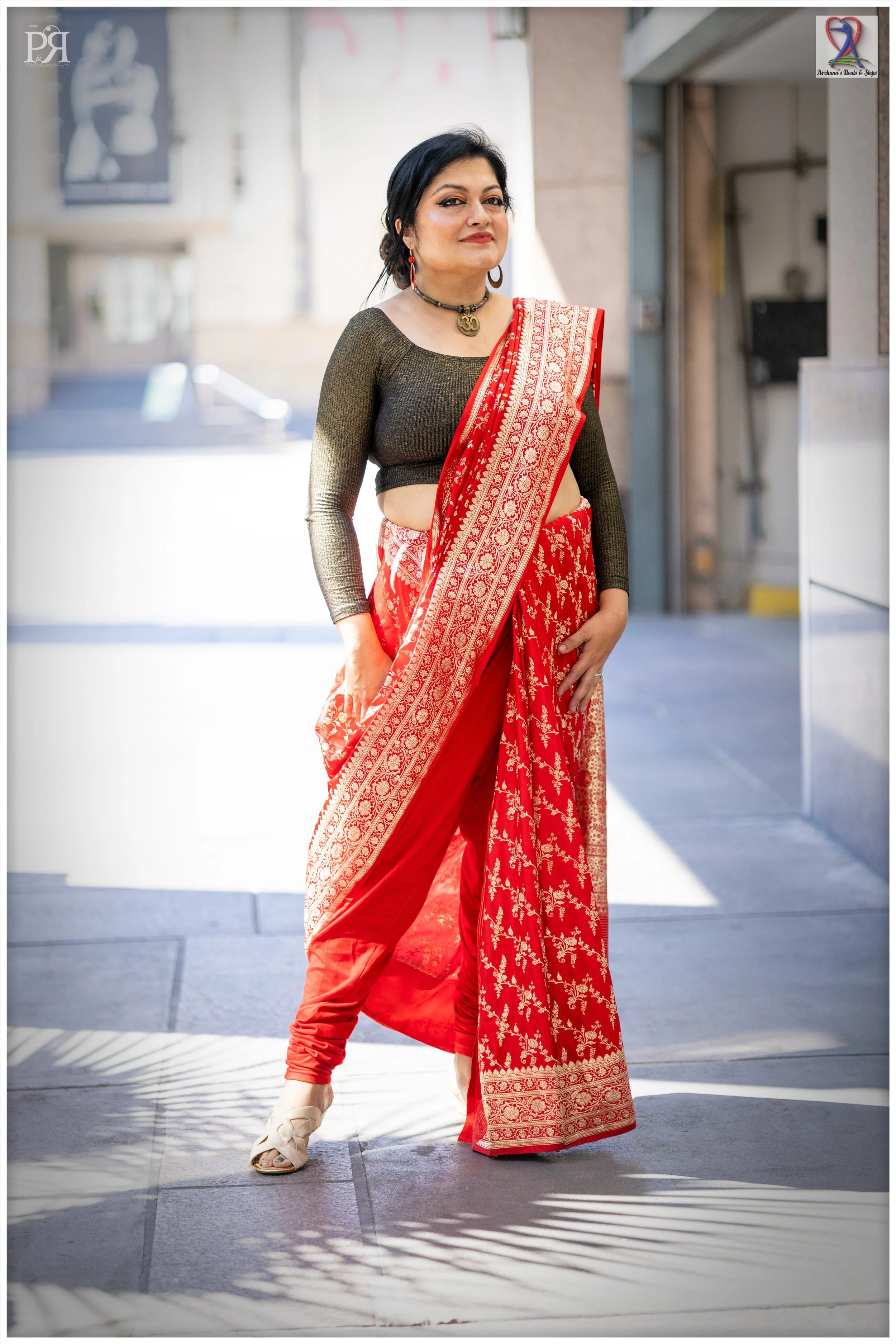 A woman wearing a red embroidered sari and a black fitted blouse, standing outdoors in sunlight.
