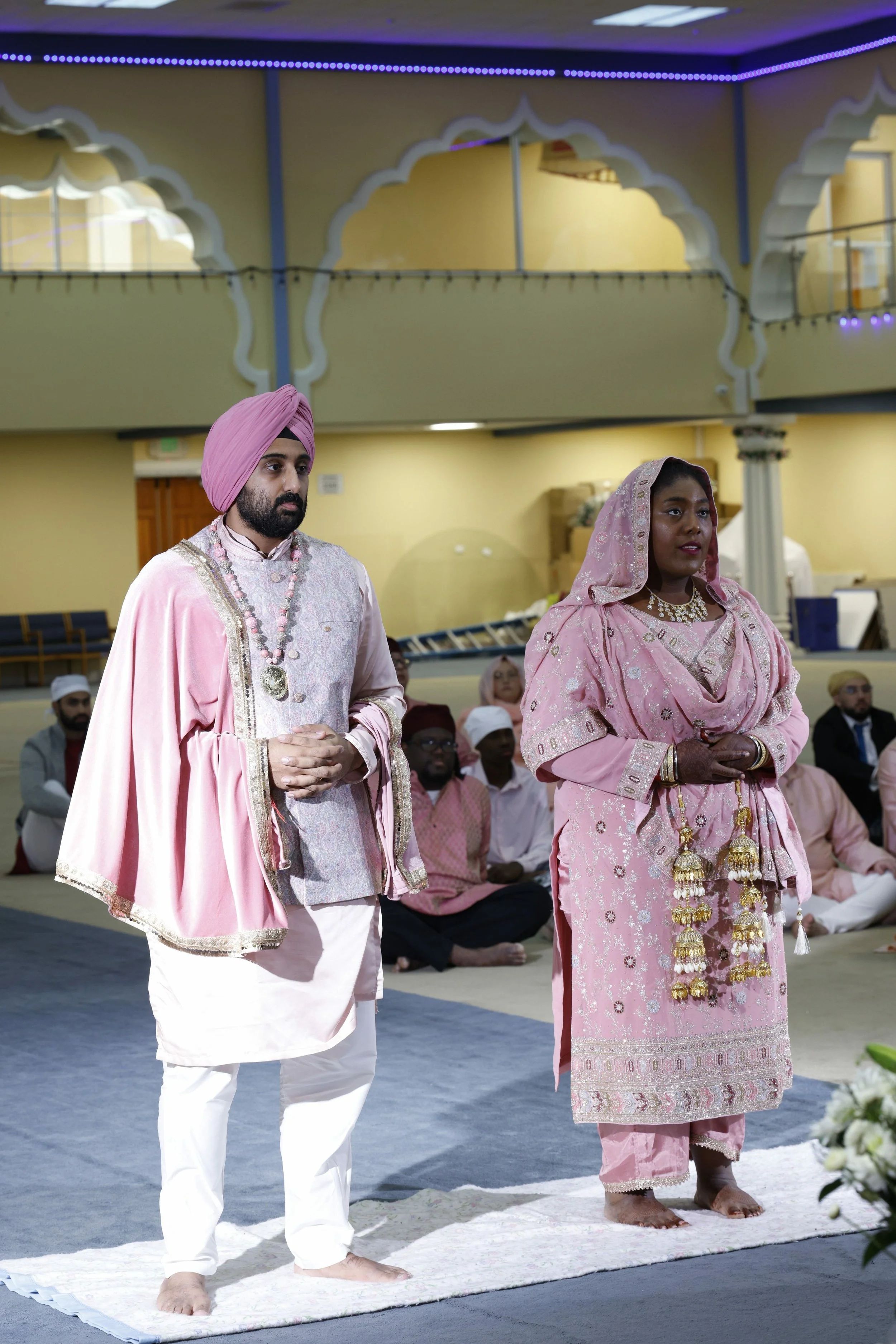 A man and woman in traditional Indian attire standing barefoot on a rug during a ceremony in a decorated hall with seated guests in the background.