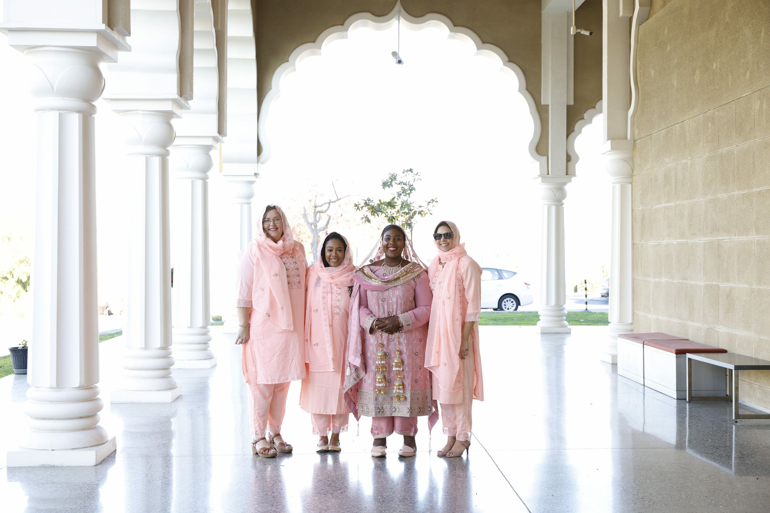 Group of women in traditional pink attire standing together outside an architectural building with arched entrance and columns.