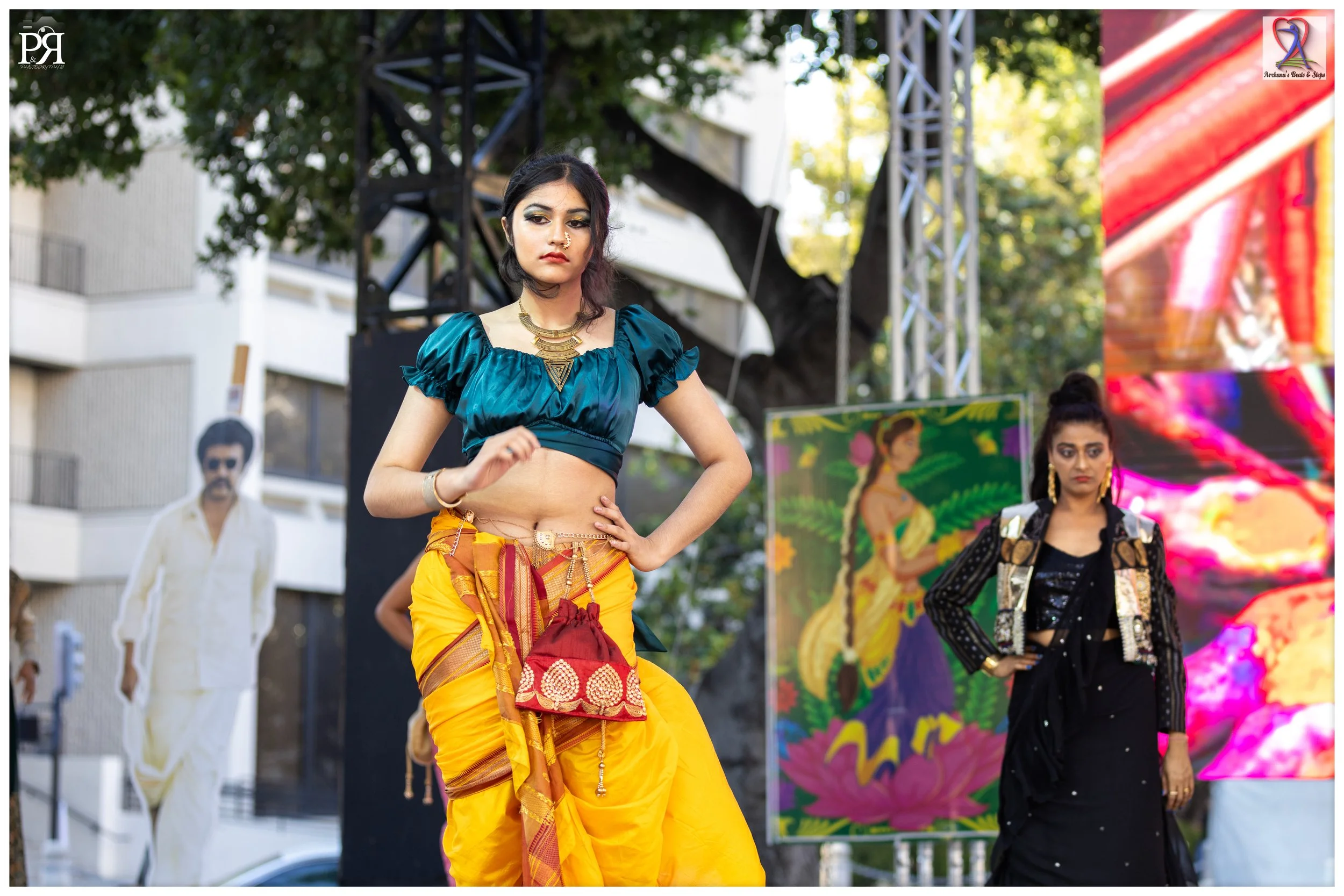 Two women standing on an outdoor stage, with one woman in traditional yellow and teal attire and jewelry in the foreground and another woman in black and gold outfit in the background, and a large digital screen displaying colorful abstract visuals.