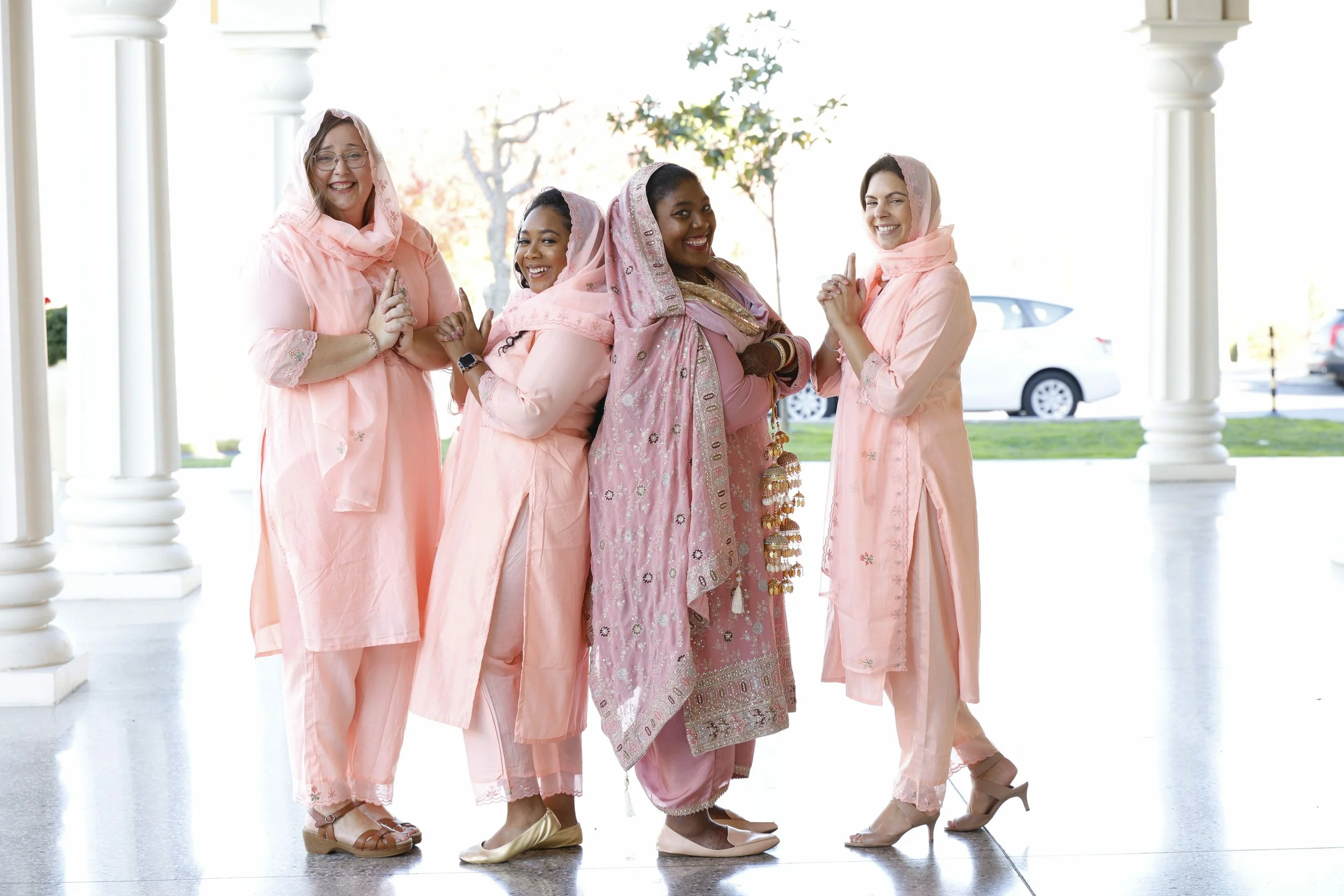 Group of five women in traditional South Asian attire, smiling and posing with hands together, outside on a porch with white columns, cars in the background.
