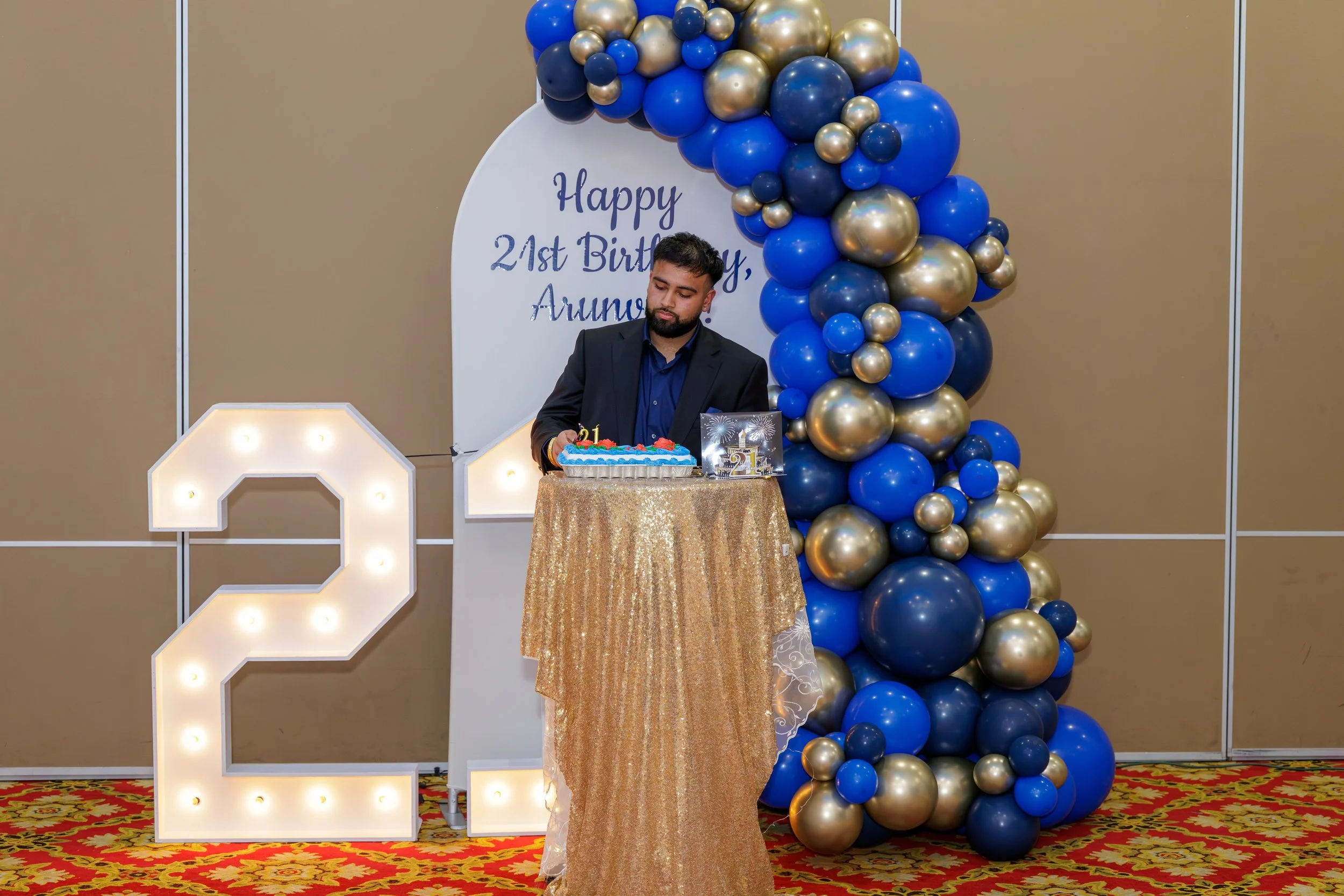 A man in a black suit and blue shirt is cutting a birthday cake at a celebration. Behind him is a white heart-shaped backdrop with blue text that reads, 'Happy 21st Birthday, Arunj.' To his left is a large illuminated number '2', and to his right is 