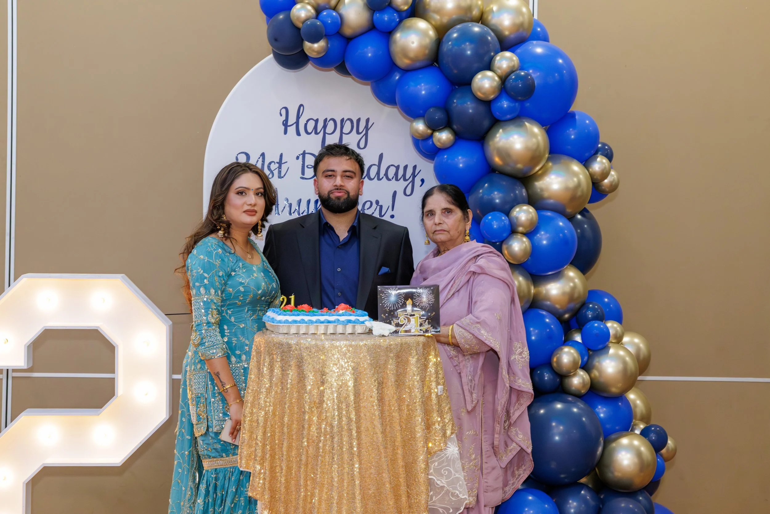 A family of three celebrating a 21st birthday with a cake, balloons, and a backdrop that reads 'Happy 21st Birthday, Mumma!'