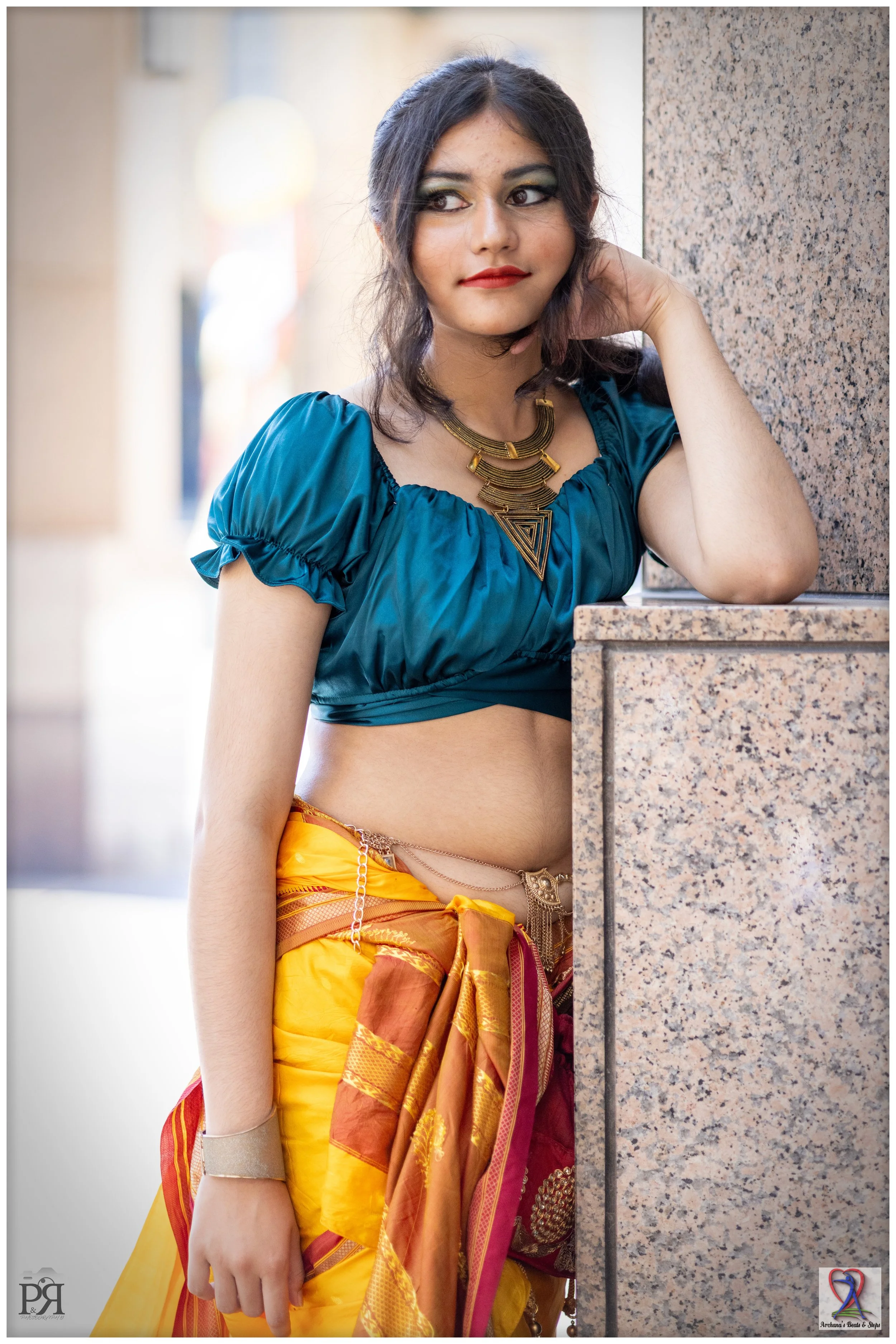 A young woman with dark hair, wearing traditional Indian attire, including a blue crop top and a yellow-orange sari with intricate patterns, leaning against a granite wall. She has bold makeup and gold jewelry, and appears outdoors with a blurred bac