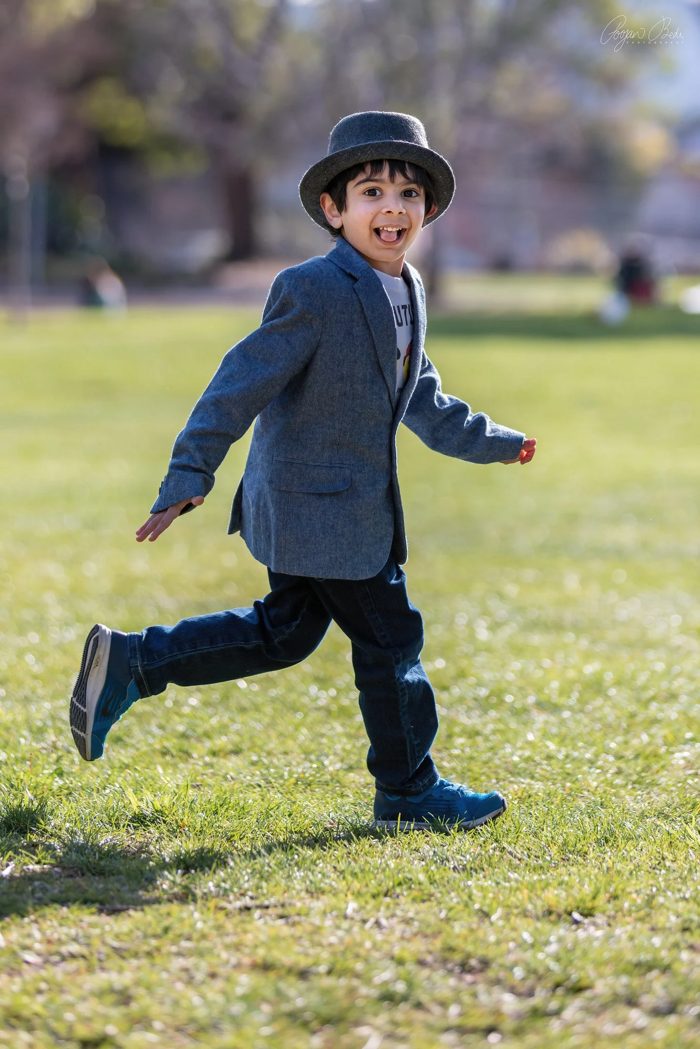 Young boy in a gray blazer and hat smiling and running on a grassy field outdoors.