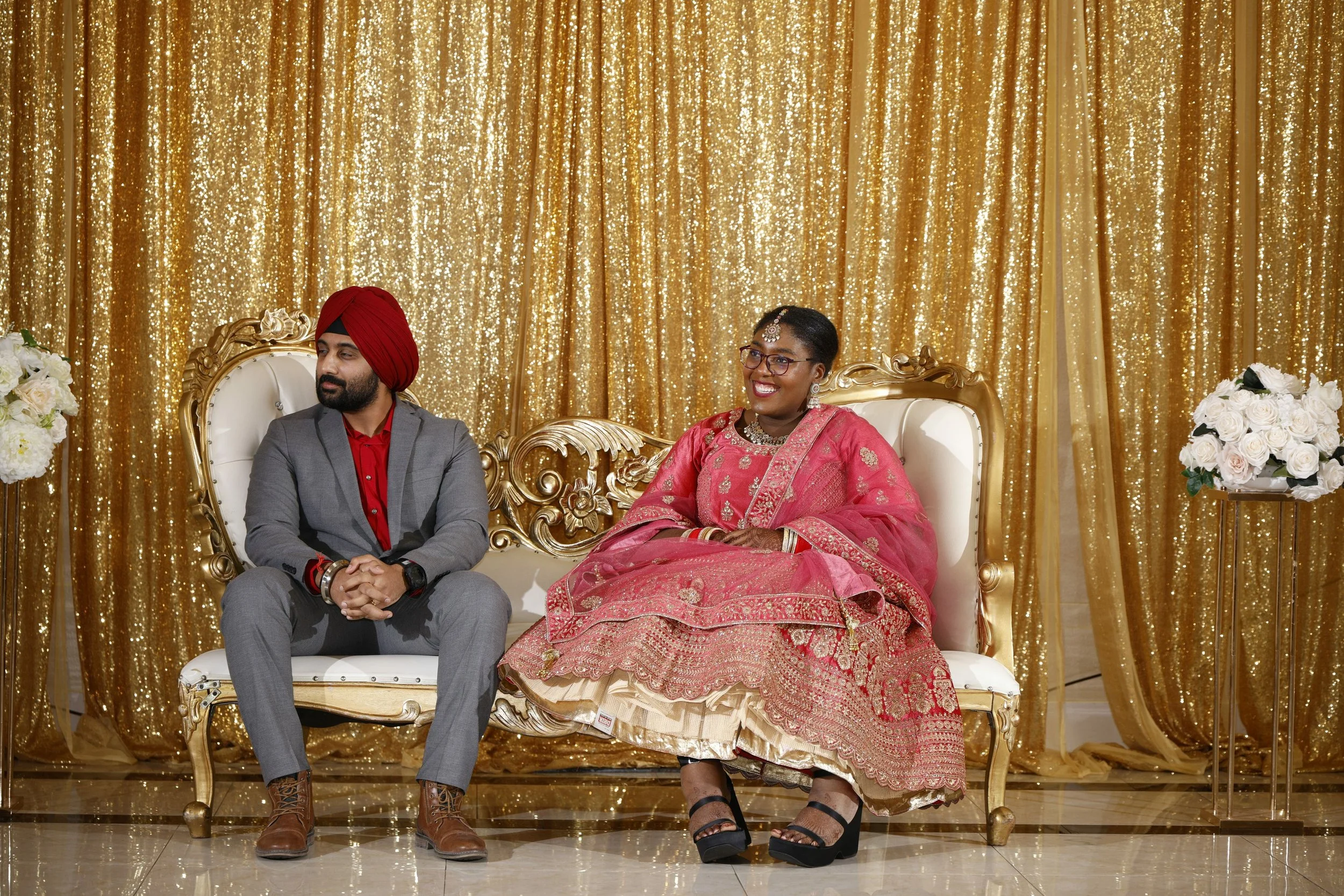 Indian couple sitting on a gold ornate couch in front of a golden sequined backdrop, with floral arrangements on either side, at a celebratory event.