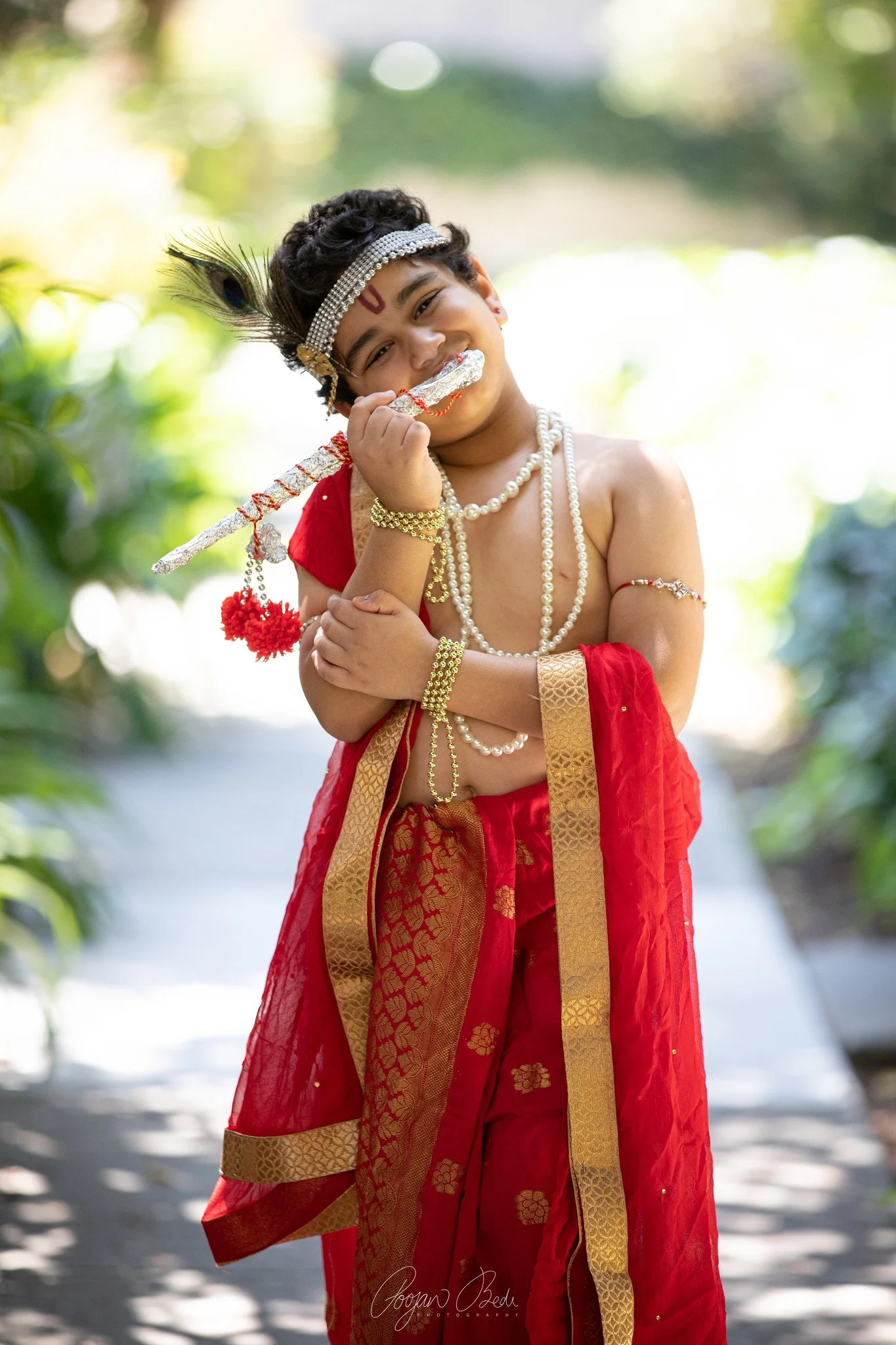 A young child dressed as Lord Krishna, wearing traditional Hindu attire with jewelry and a peacock feather in his hair, holding a flute and smiling outdoors.