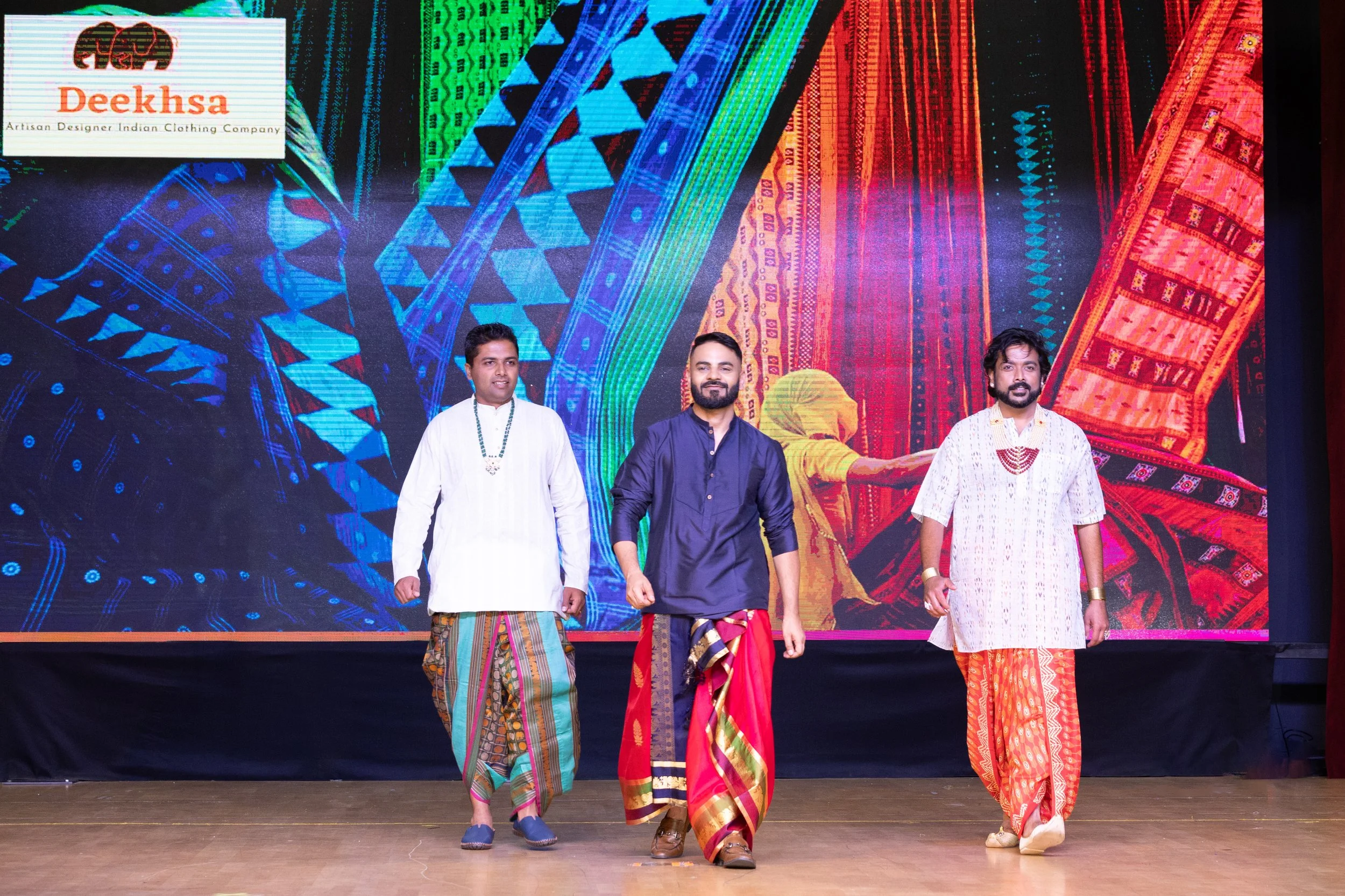Three men walking on stage in traditional Indian attire, with a colorful digital backdrop and a logo in the upper left corner.