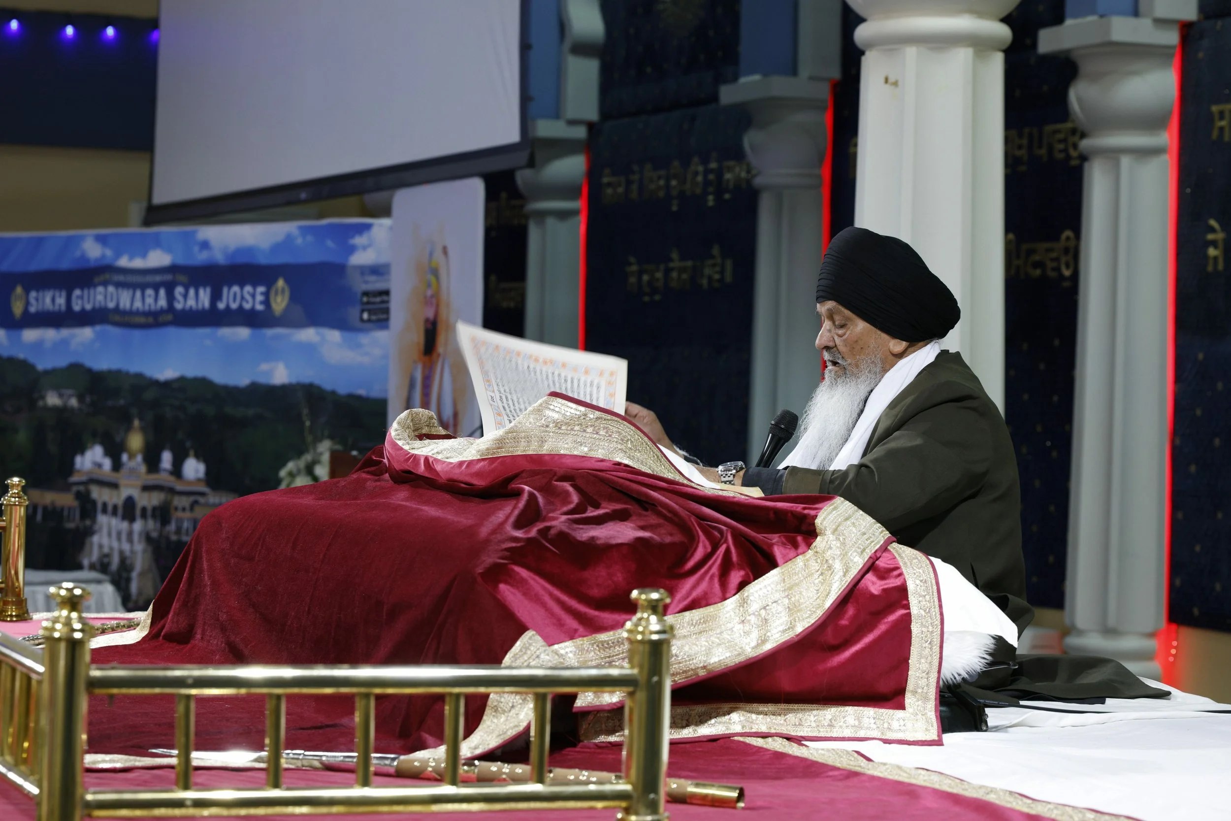 An elderly Sikh man with a long white beard, wearing a black turban and a white scarf, sitting cross-legged and reading from a religious text on a raised platform with a red cover, in a Sikh temple with decorated columns and a banner that reads 'Sikh