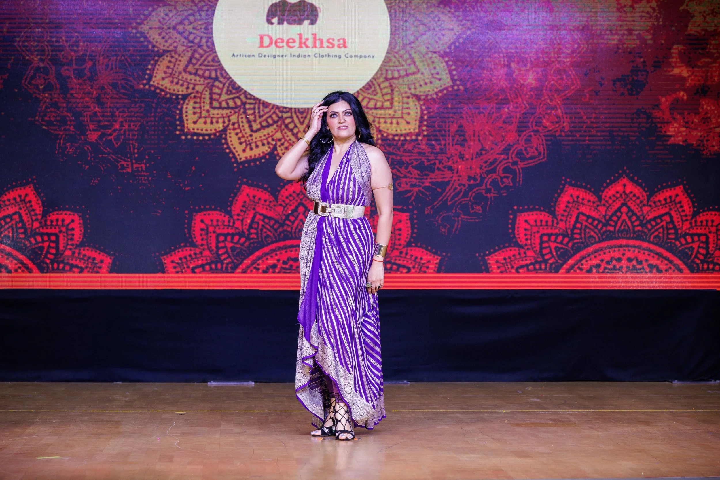 A woman in a purple and white saree walking on stage during a fashion event with a backdrop that reads "Deeksha" and features a decorative mandala pattern.