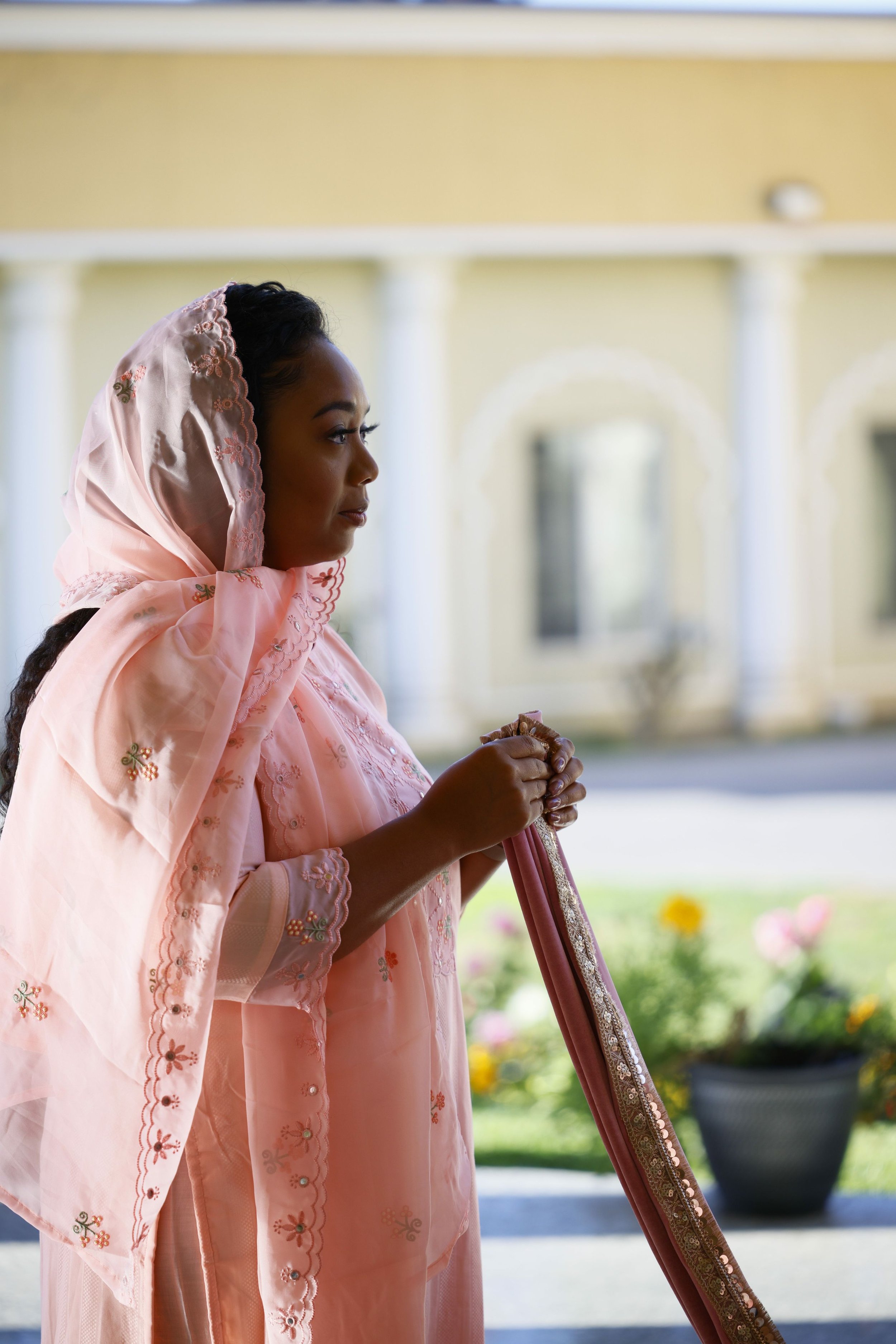 A woman dressed in traditional South Asian attire, holding a decorative cloth, standing outdoors in front of a building with arched windows and potted plants.