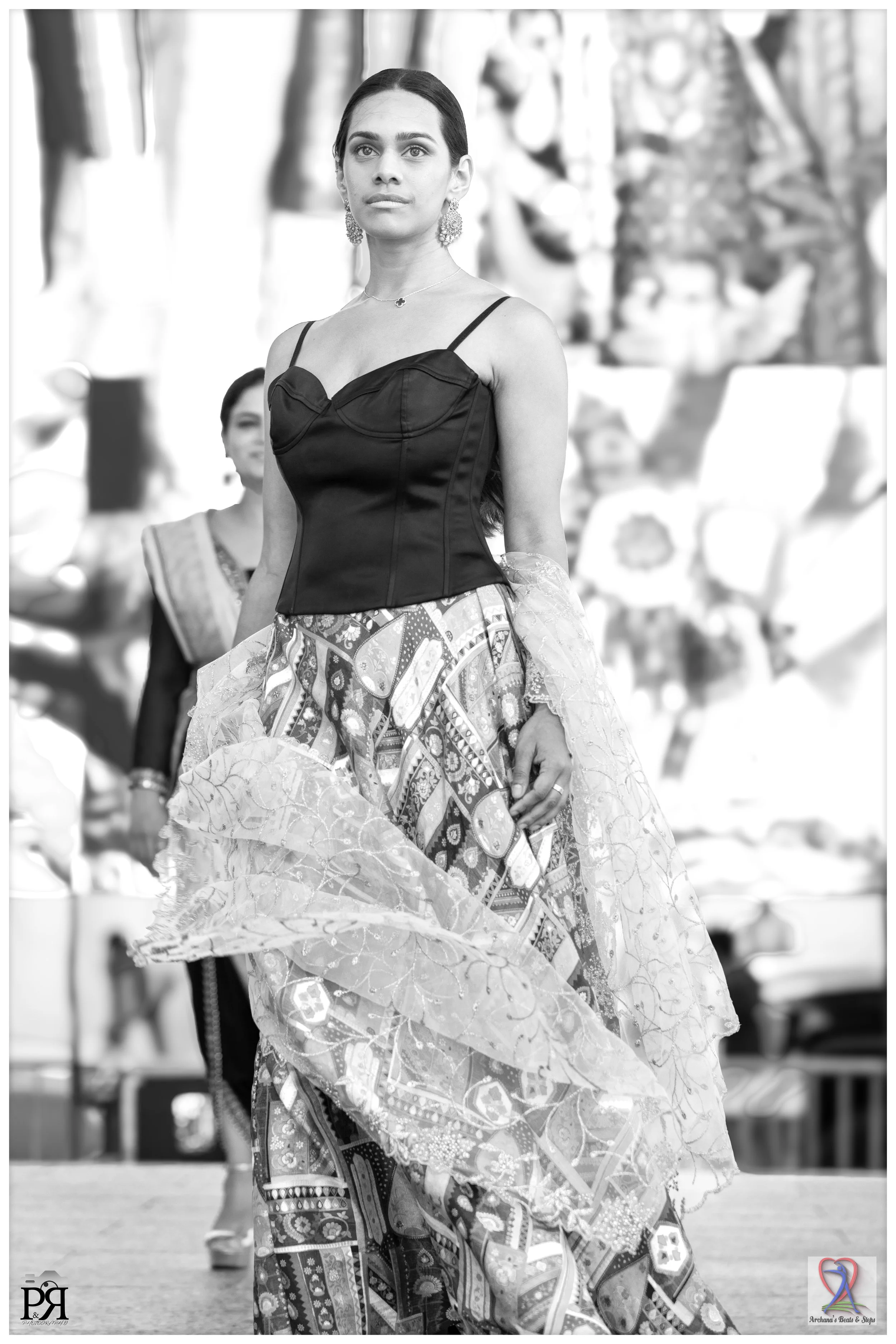 Woman walking on a runway in traditional Indian attire, wearing a sleeveless black top and a flowing, patterned skirt with sheer overlay.