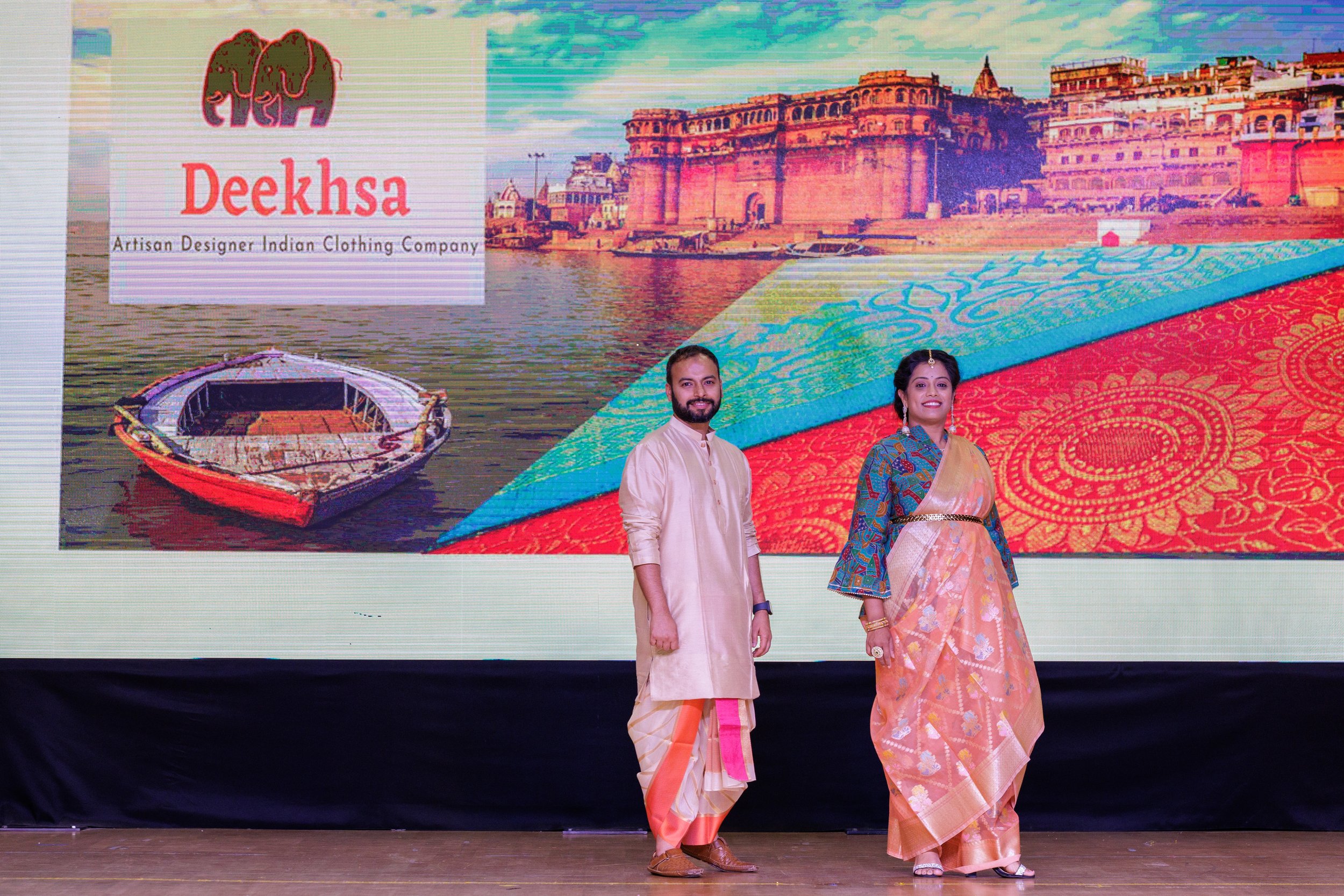 Two people dressed in traditional Indian attire standing on stage in front of a large screen displaying the logo and name 'Deeksha, Artisan Designer Indian Clothing Company,' along with an image of a boat on the river and historic Indian architecture
