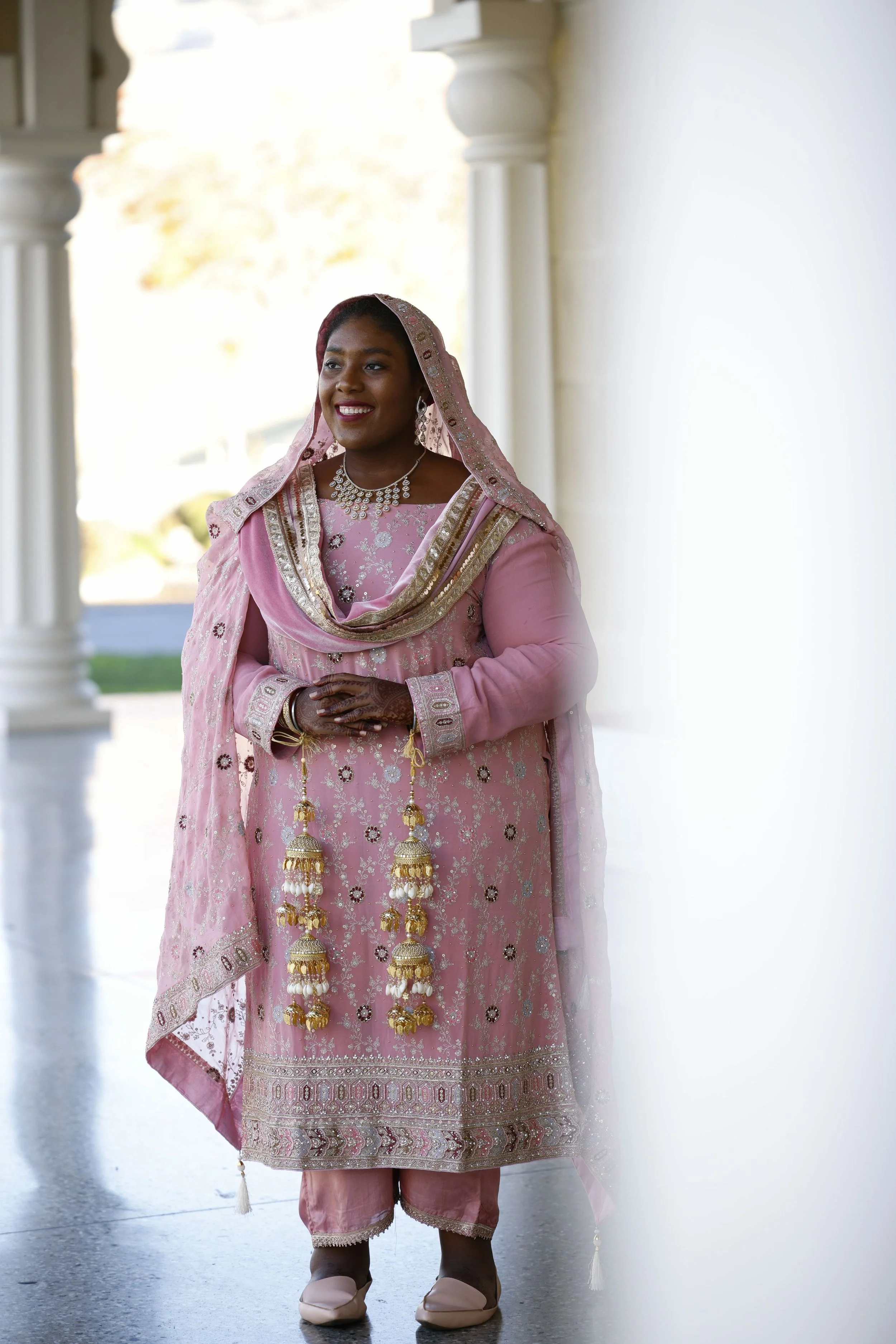 A woman dressed in traditional pink Indian attire with jewelry, standing near white columns and smiling.