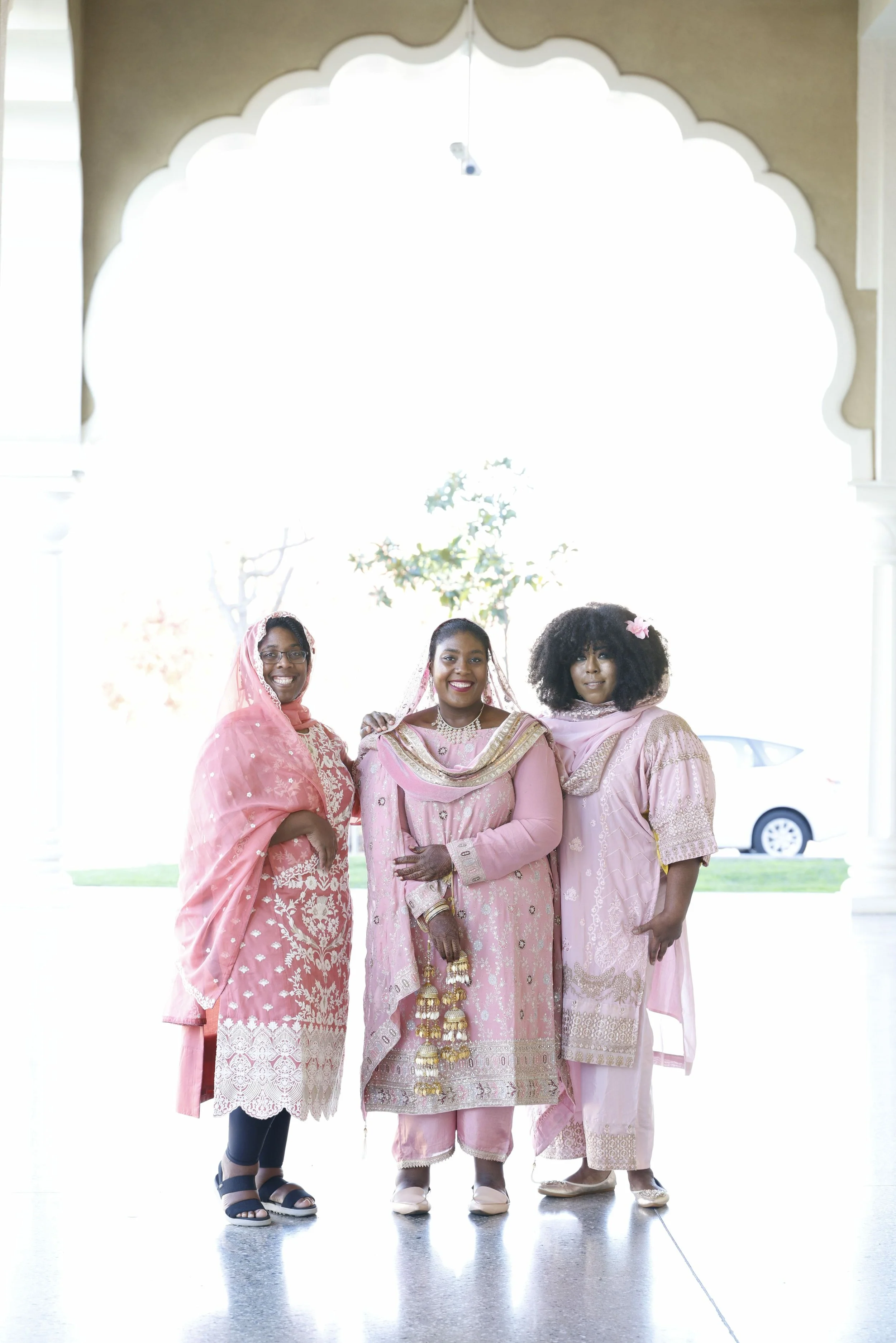 Three women dressed in traditional pink and gold Indian attire, standing together inside a bright building with an ornate archway in the background.