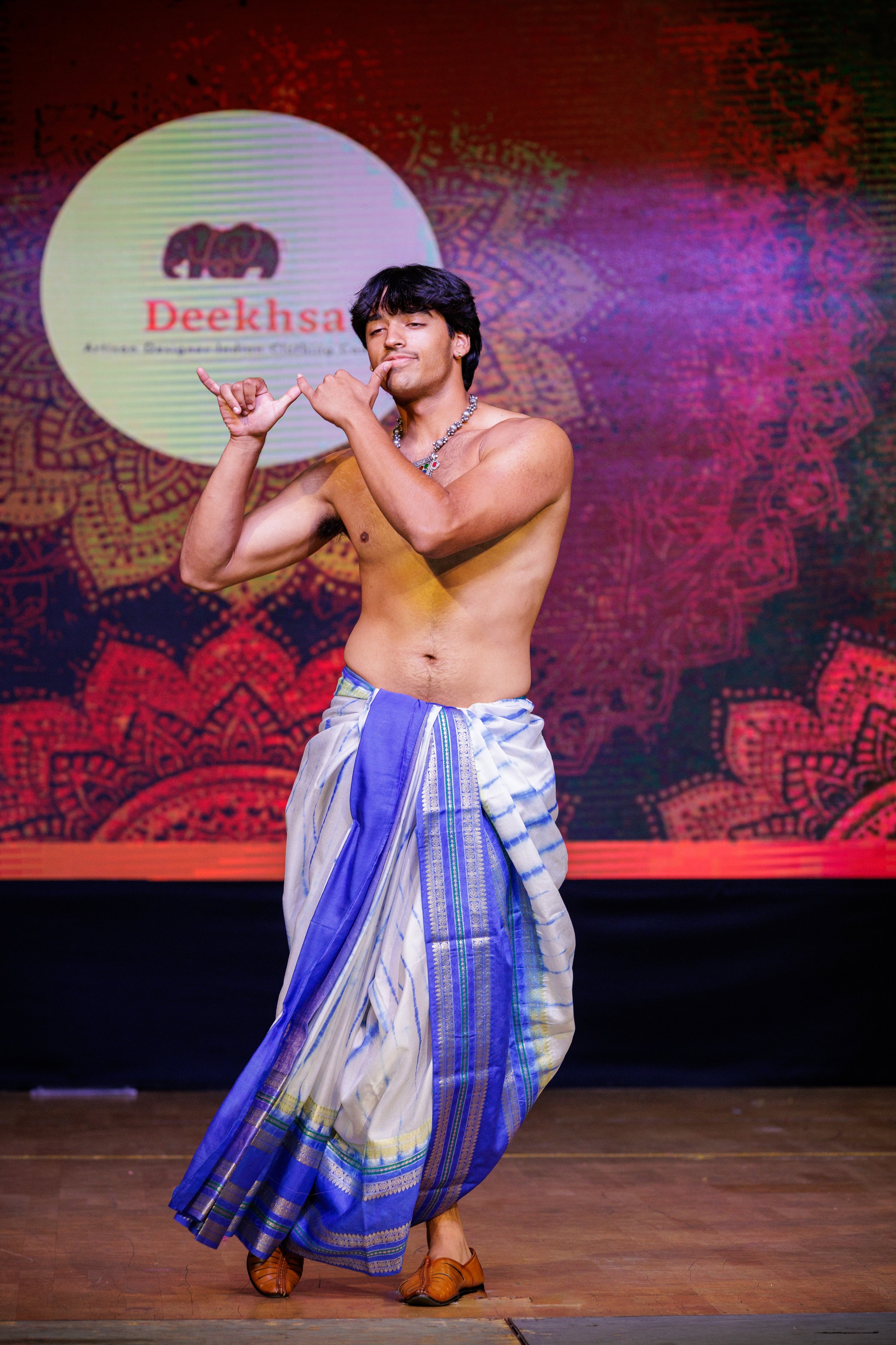 A shirtless man wearing traditional South Indian dhoti looks at the camera while striking a dance pose on stage with a colorful backdrop that has a logo and the word "Deeksha."