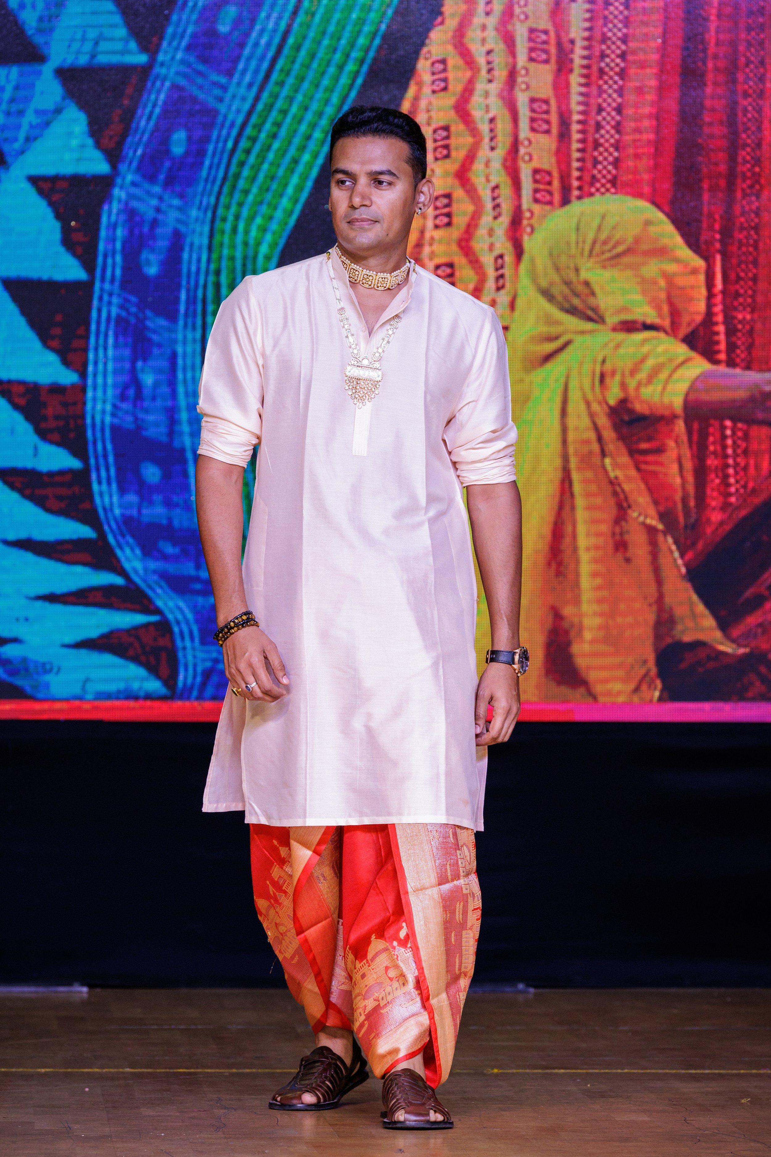 A man dressed in traditional Indian attire walking on a stage with a colorful patterned background.