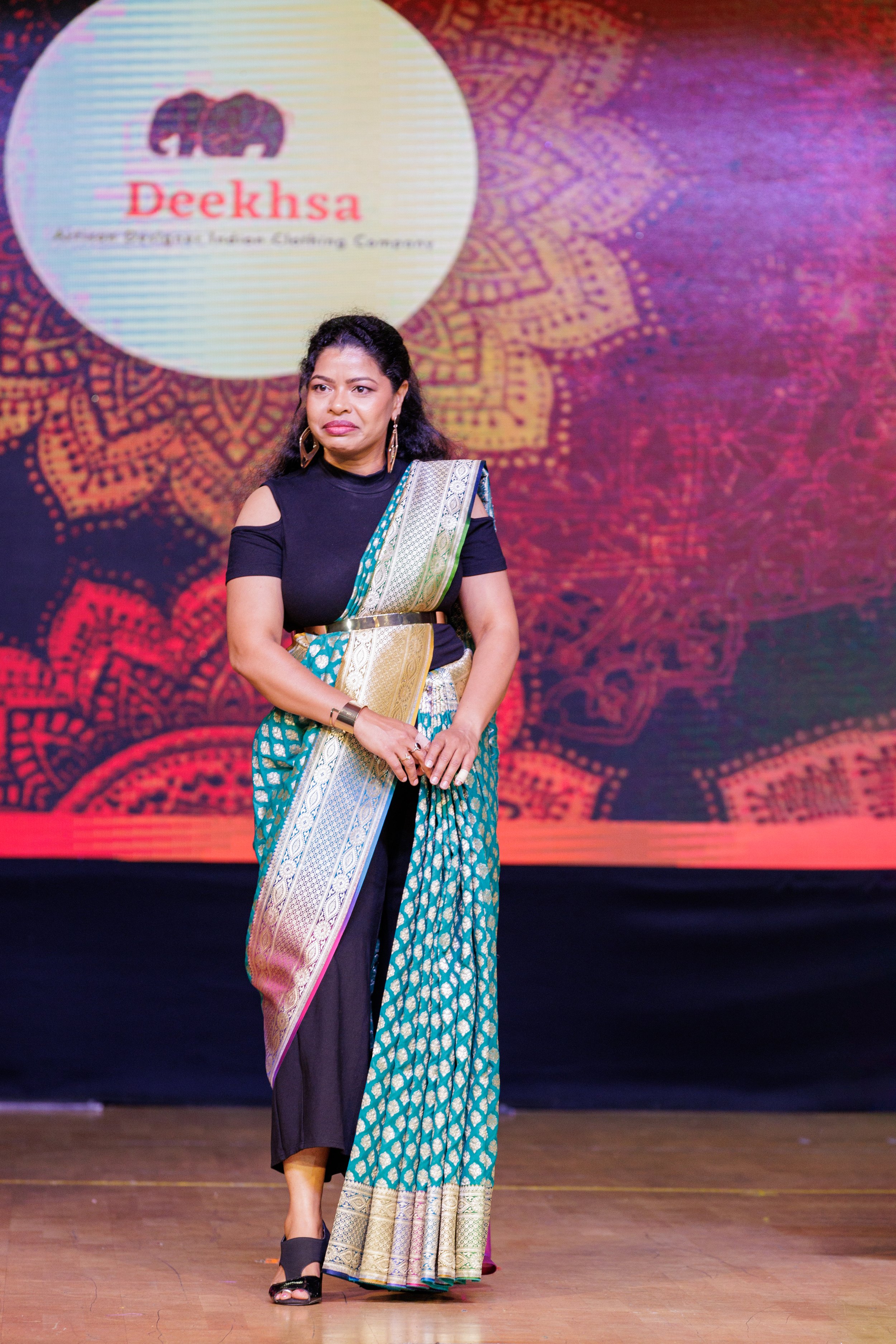 A woman in a traditional Indian sari standing on a stage with a colorful background and a logo that reads 'Deeksha'.