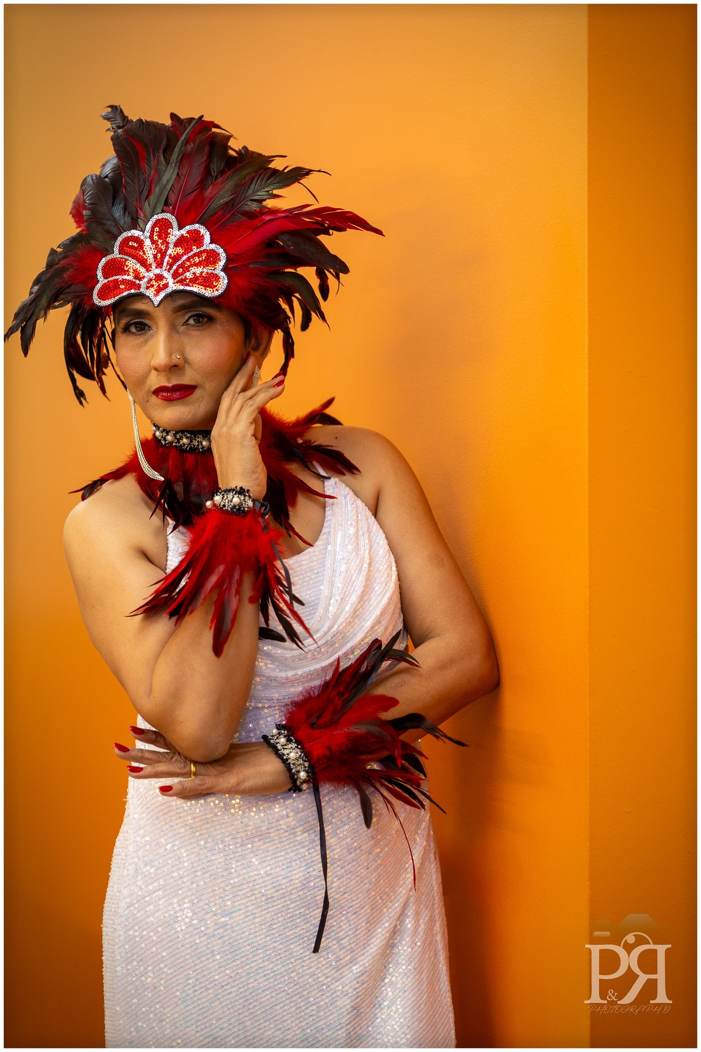 Woman in a white dress with red and black feathered accessories and a feathered headpiece, standing against an orange background.