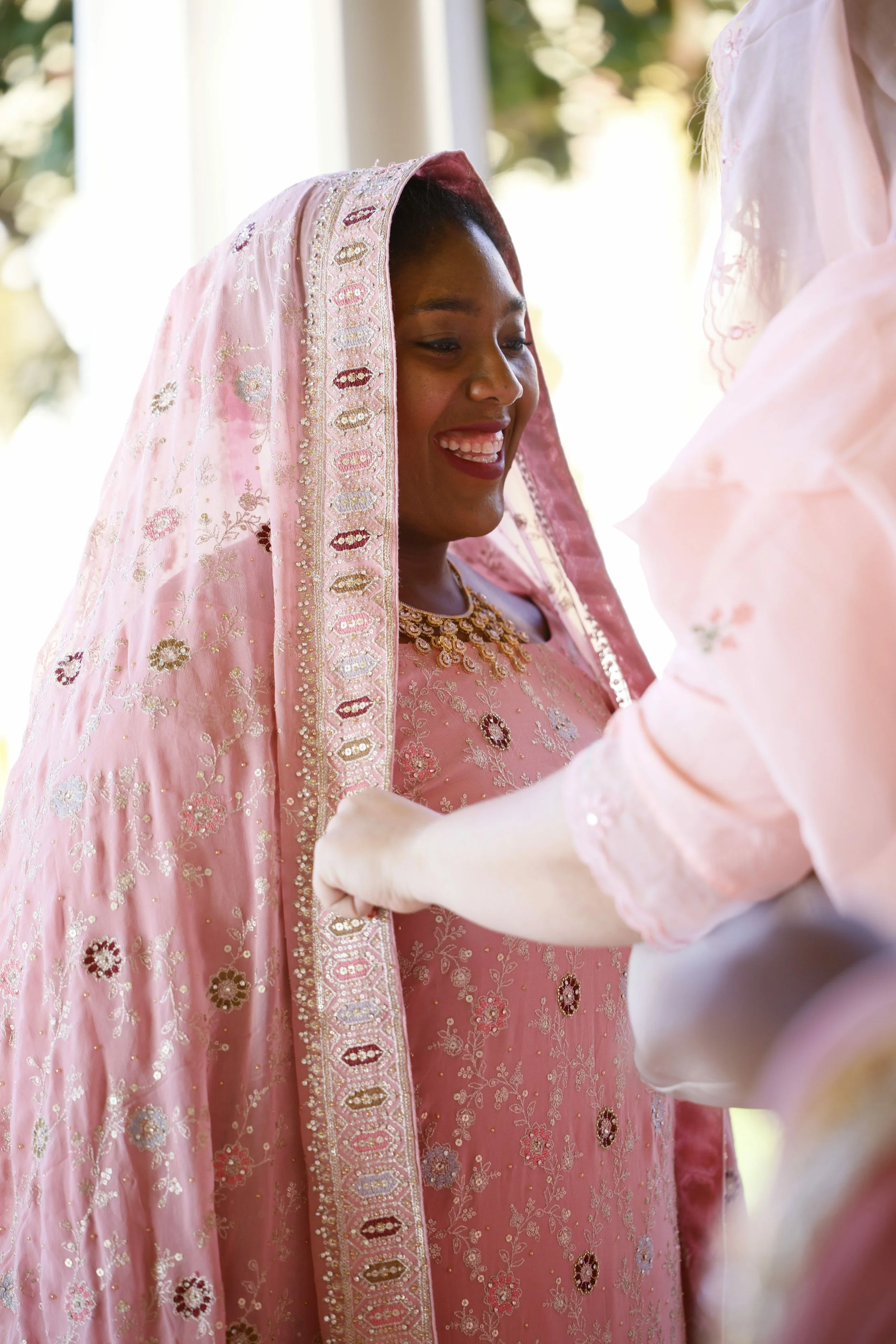 A woman in traditional pink embroidered attire with a matching headscarf and jewelry, smiling during a cultural or ceremonial event.
