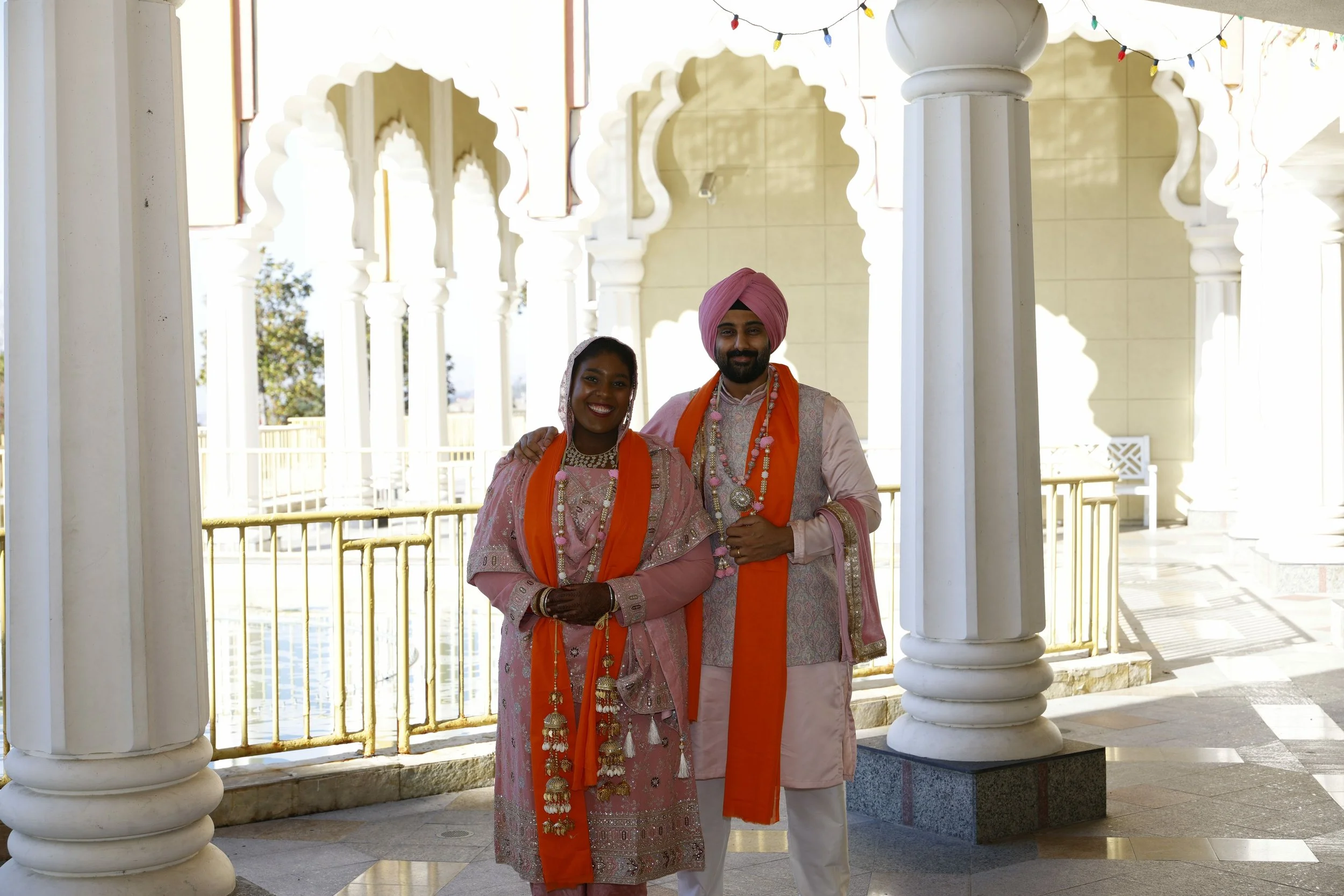A couple dressed in traditional Indian wedding attire standing together in an ornate architectural setting with white columns and arches, decorated with string lights, during daytime.