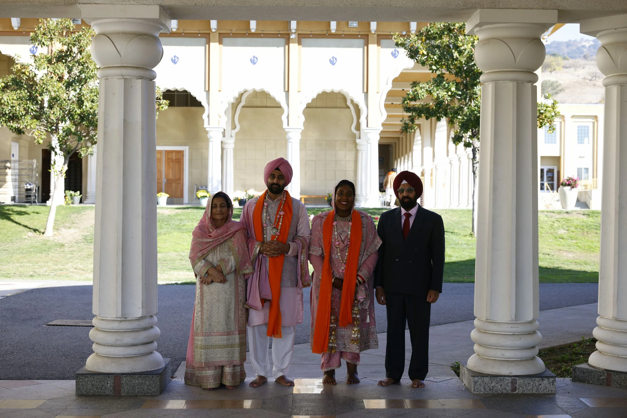 A group of five people dressed in traditional Indian attire standing outdoors beneath a white columned structure, with a building, trees, and a grassy area in the background.