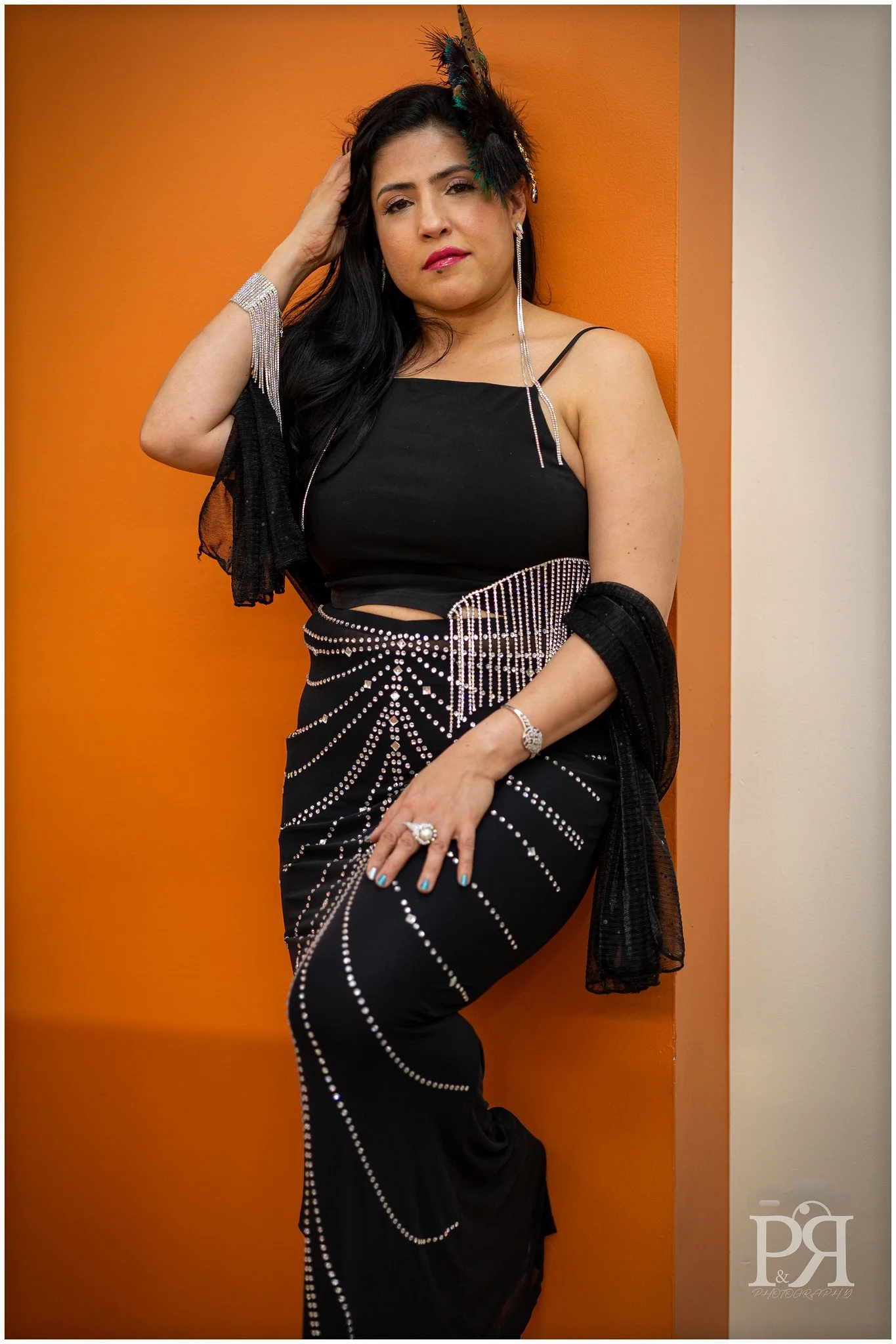 A woman with long dark hair in a black dress with rhinestone embellishments, wearing a feathered headpiece, earrings, bracelet, and ring, leaning against an orange wall.