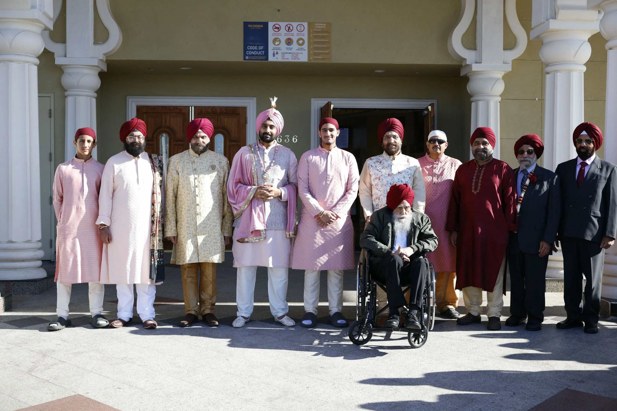 Group of men dressed in traditional Indian attire, standing outdoors in front of a building, with one man in a wheelchair.