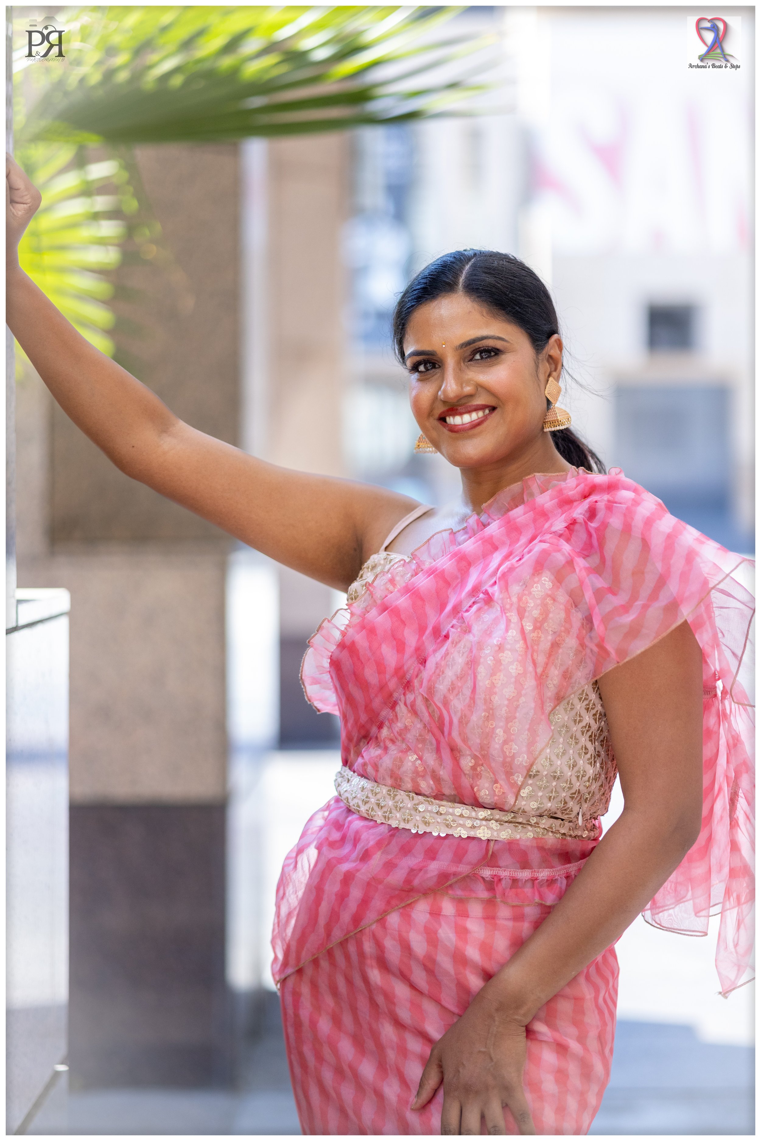 A woman wearing a pink checkered saree, gold earrings, and smiling, standing outdoors with her arm resting on a wall, with blurred buildings in the background.