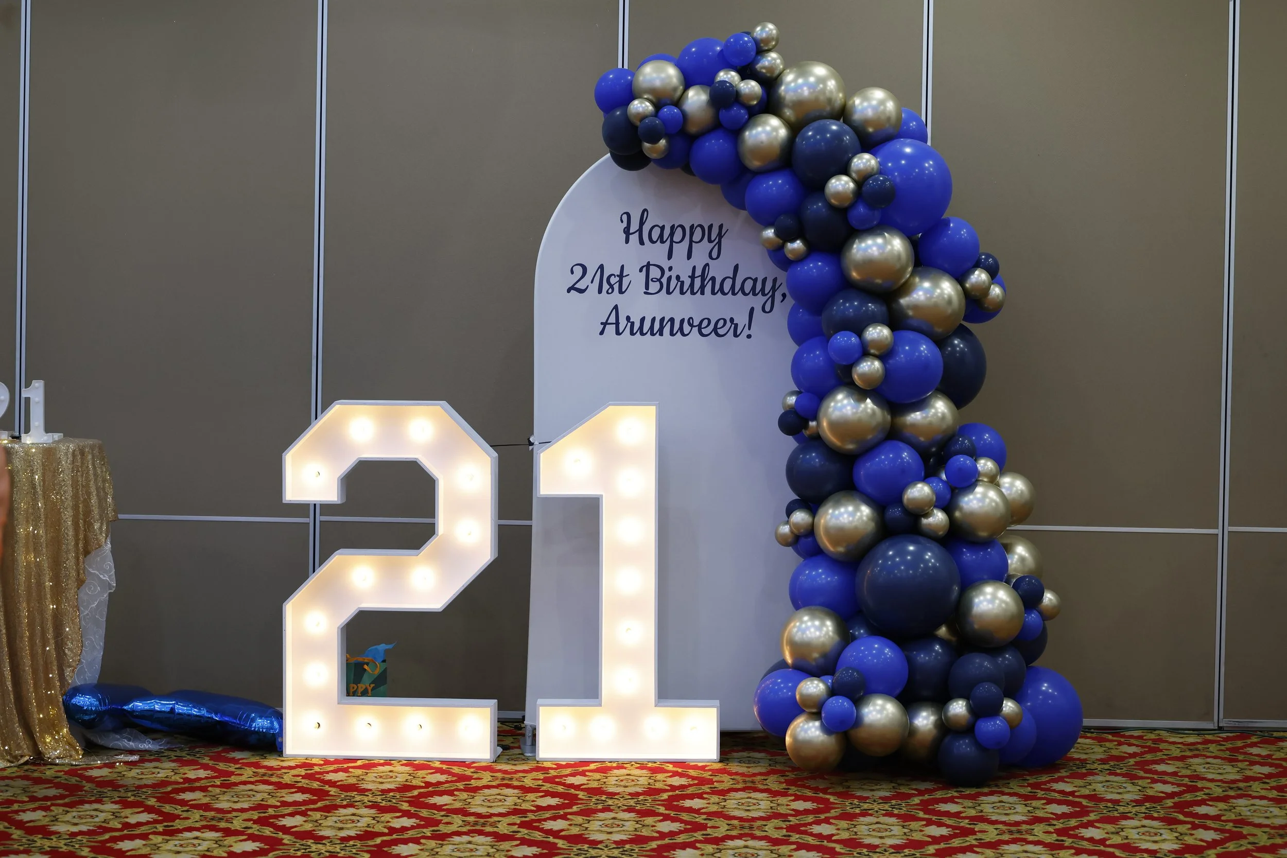 Decorative birthday setup with large illuminated '21' sign, gold and blue balloons arranged in an arch, and a sign that reads 'Happy 21st Birthday, Arunveer!' on a white backdrop.