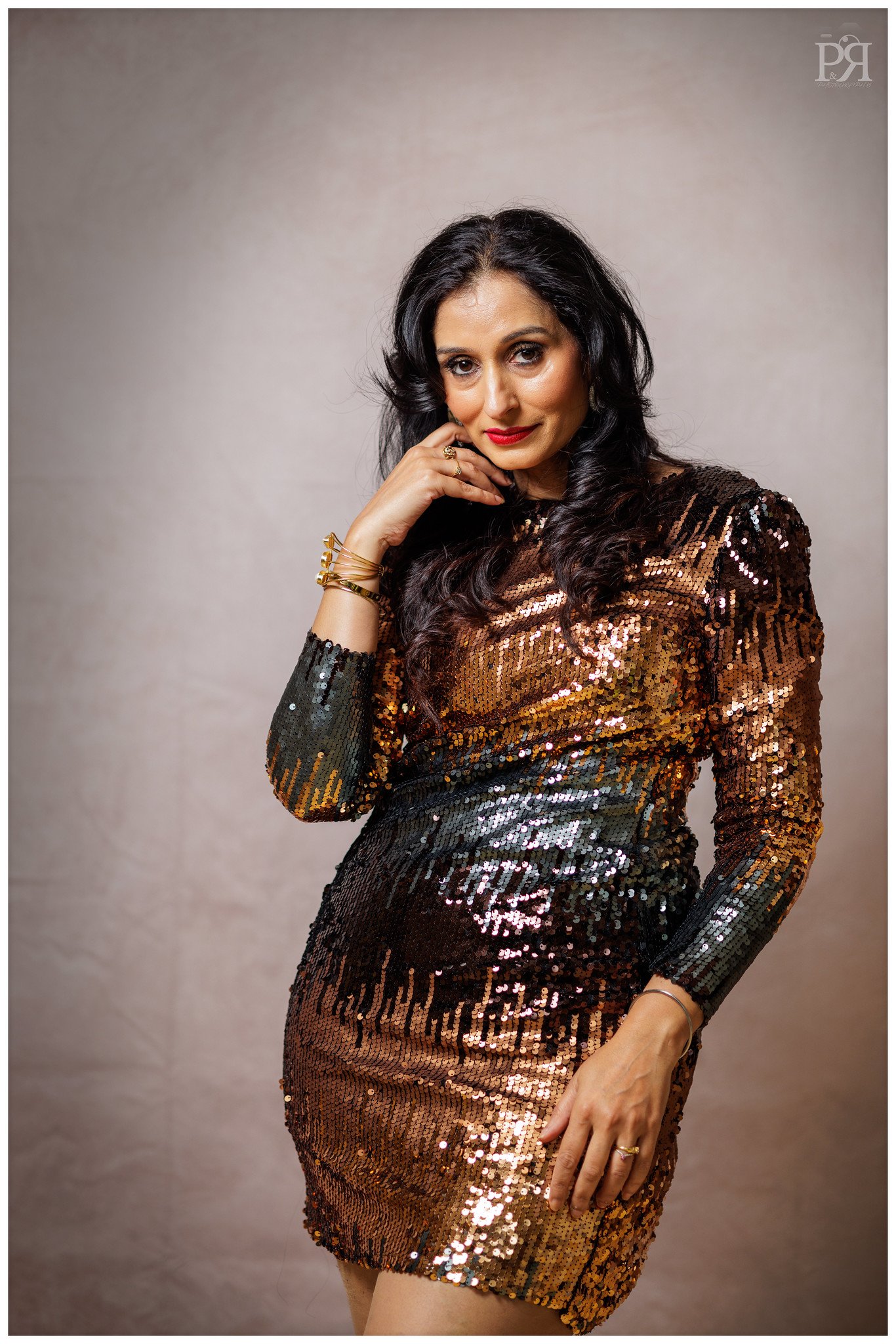 A woman with dark wavy hair wearing a shiny, sequined black and gold dress, posing with one hand near her face and the other resting on her thigh, against a neutral background.