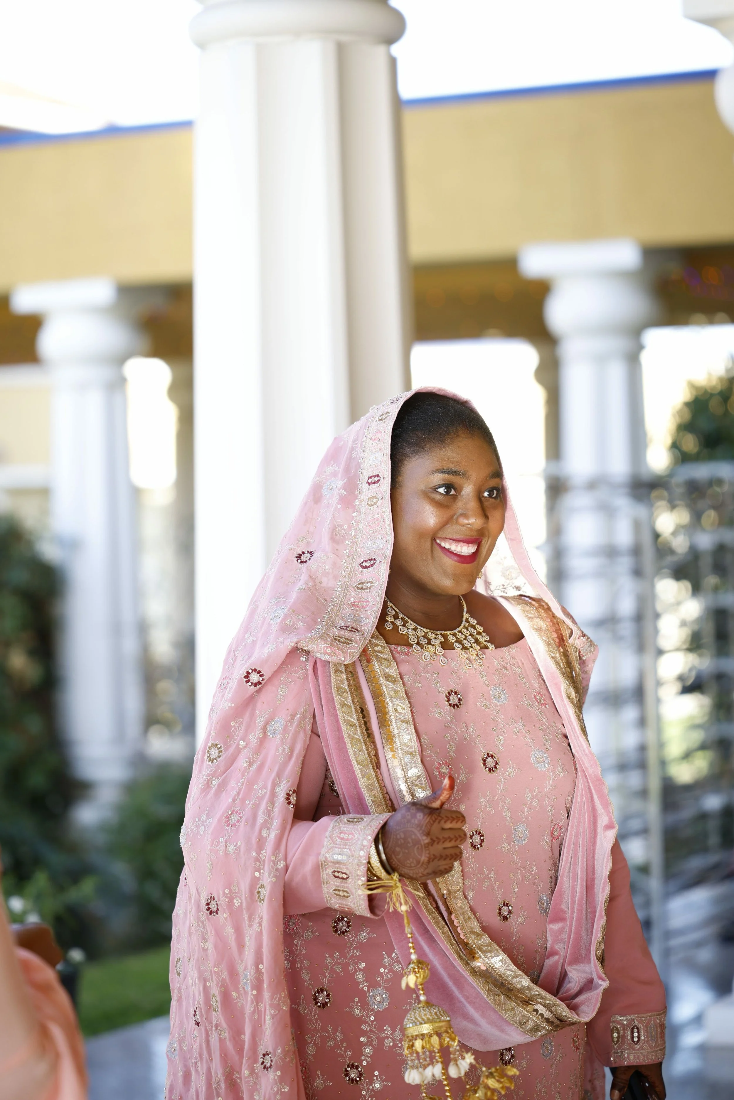 A smiling woman dressed in traditional Indian pink attire with jewelry, giving a thumbs-up gesture, standing outdoors in front of white columns and a building.